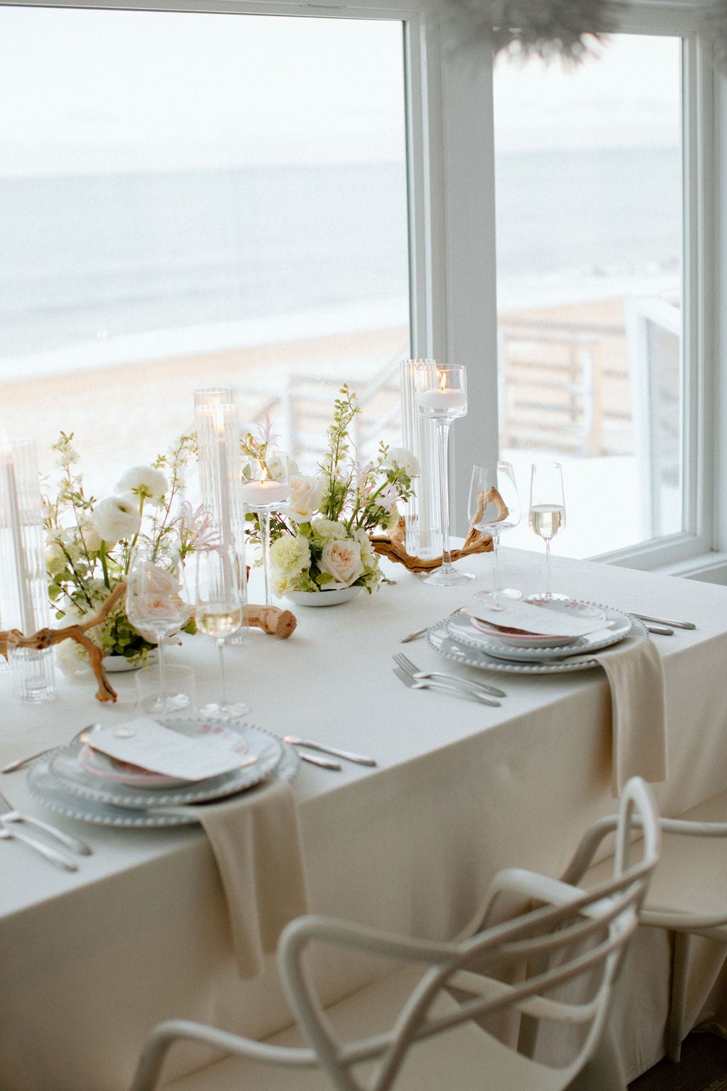 A fine dining table set with white tablecloth, flowers, candles, and wine glasses, with a view of the ocean in the background.