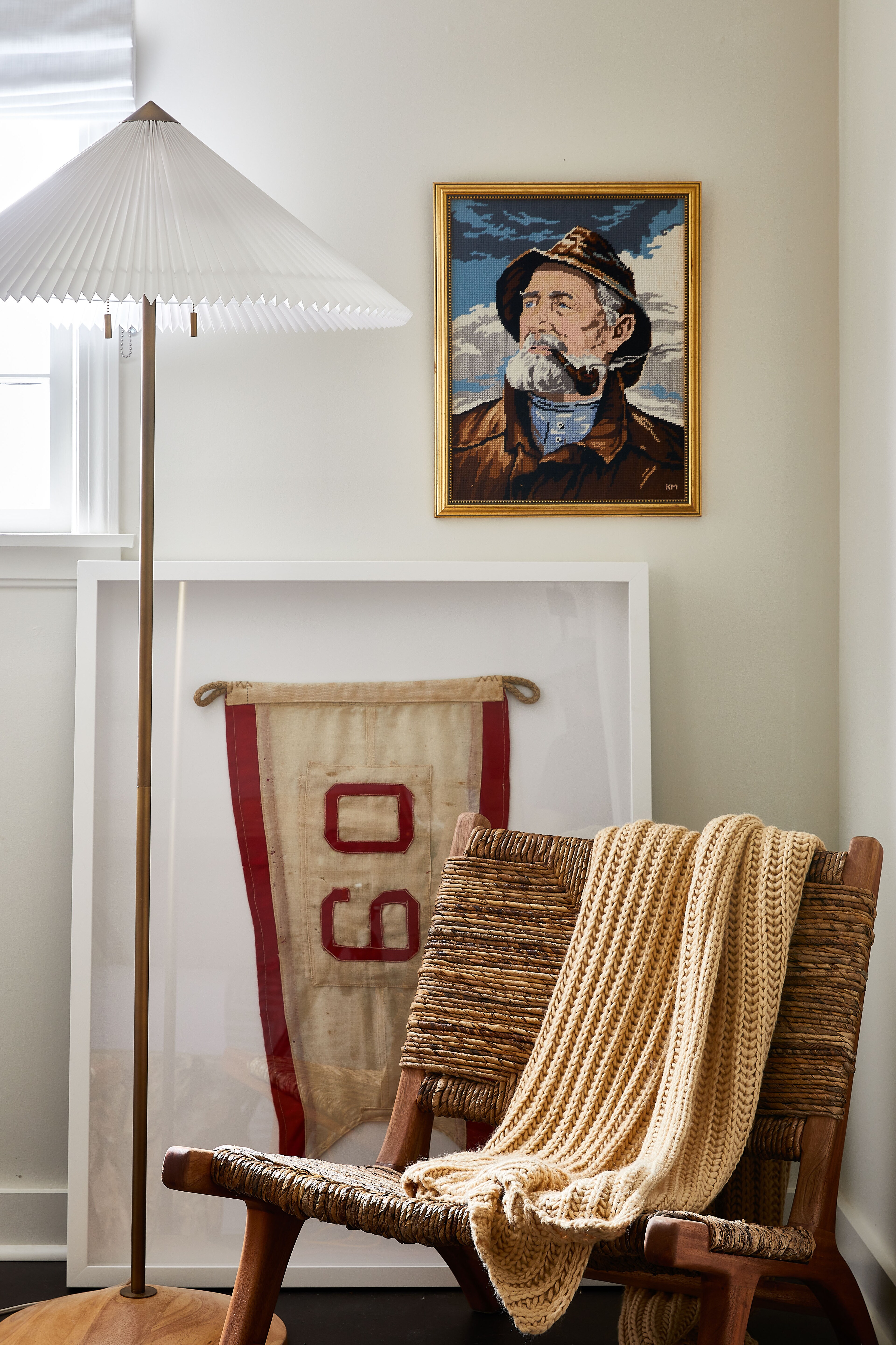 Woven lounge chair with a knit blanket beside vintage artwork and a floor lamp.