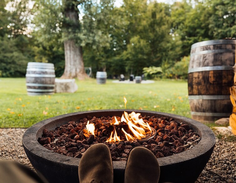 Guest relaxing beside an outdoor fire pit surrounded by trees and wooden barrels.