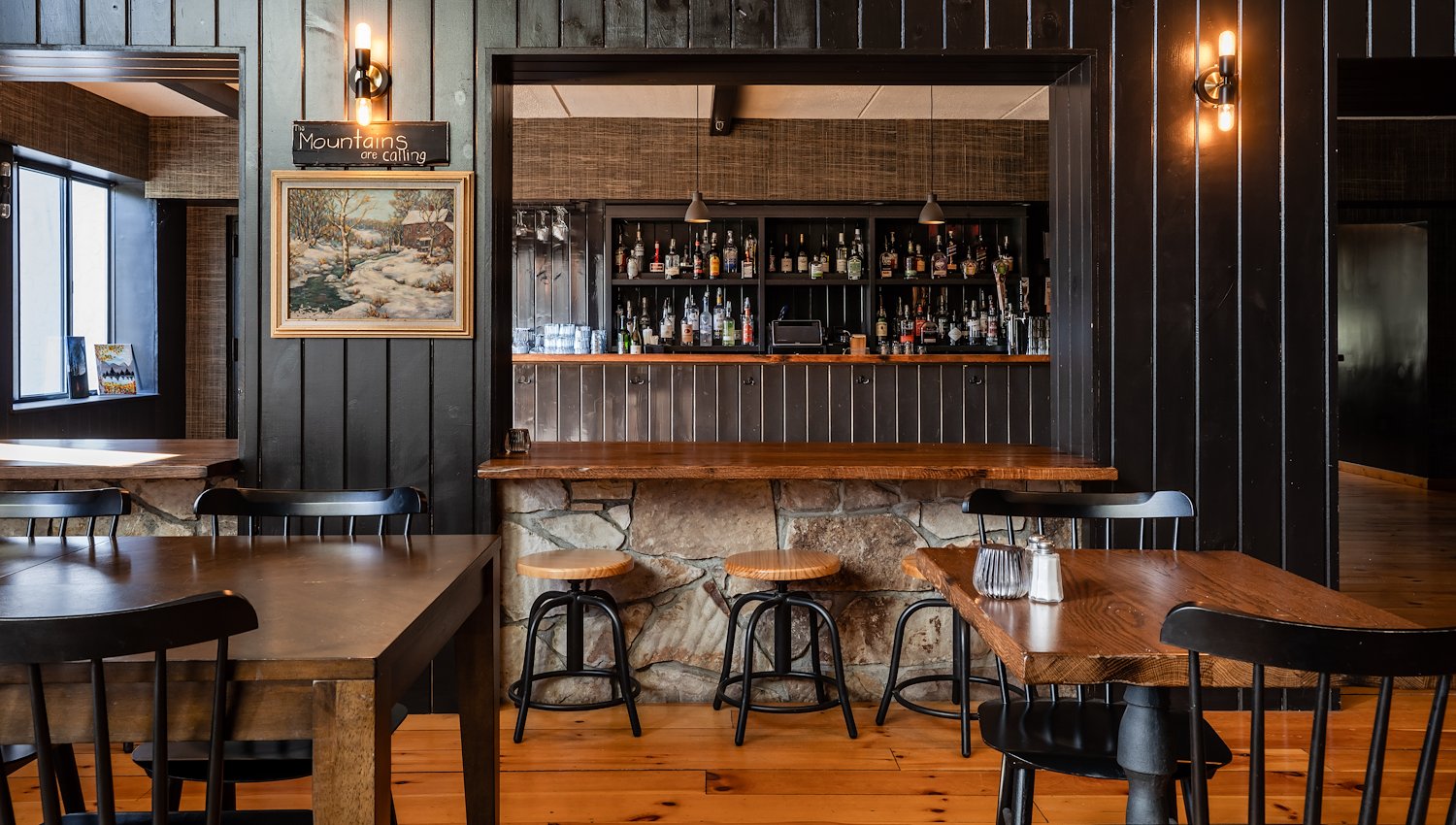 Rustic hotel bar and dining area with wood tables, stone accents and dark paneled walls.