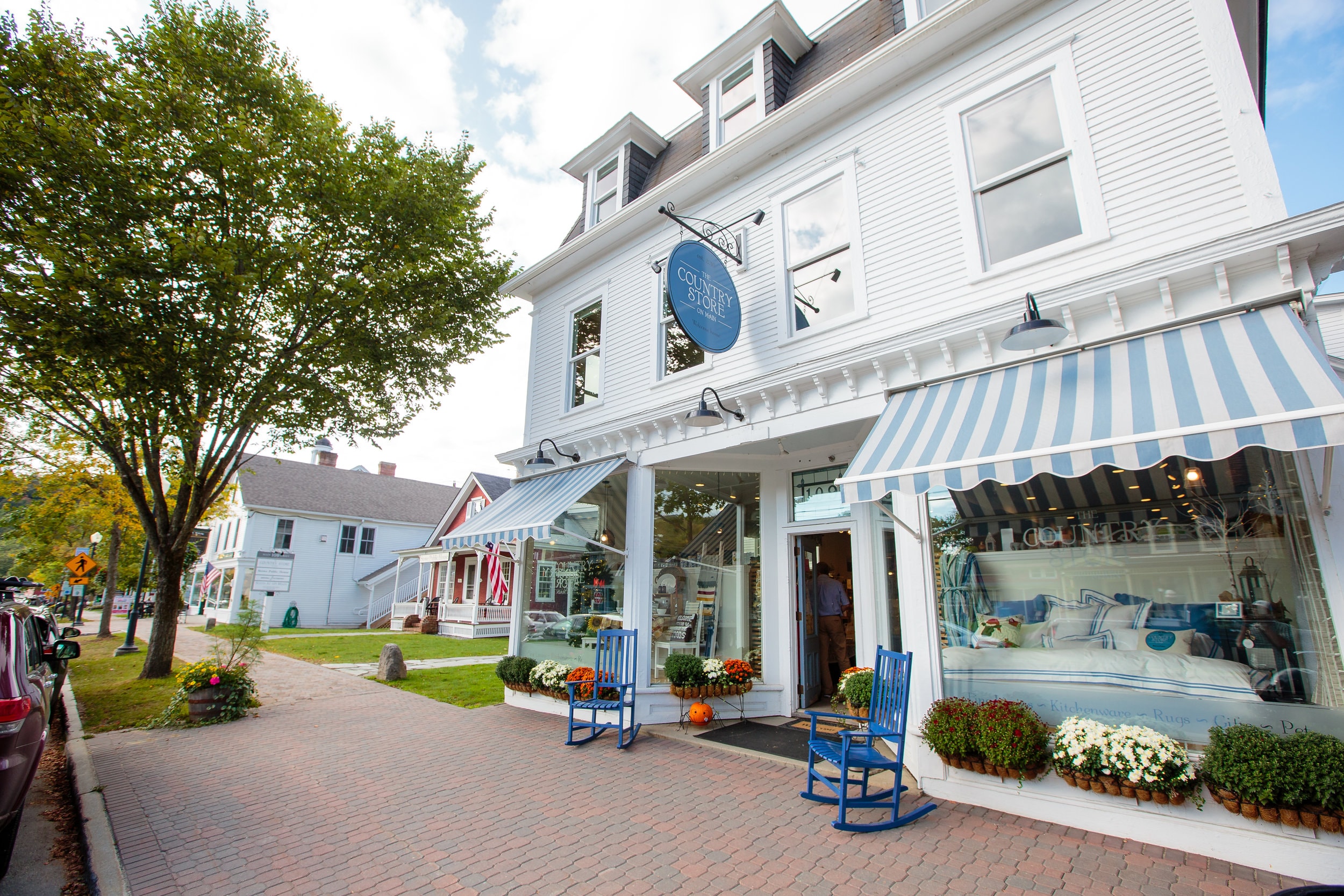 A white building with a blue and white awning and blue rocking chairs on a brick sidewalk