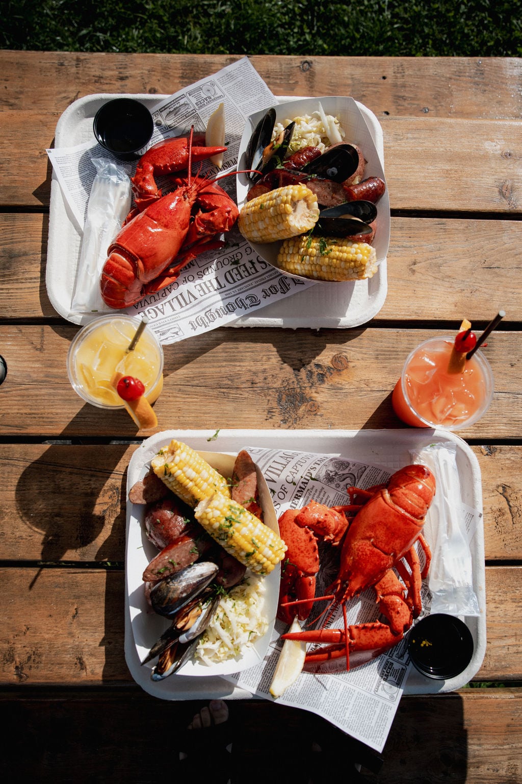 Two seafood meals on paper-lined trays with lobster, mussels, corn, and drinks.