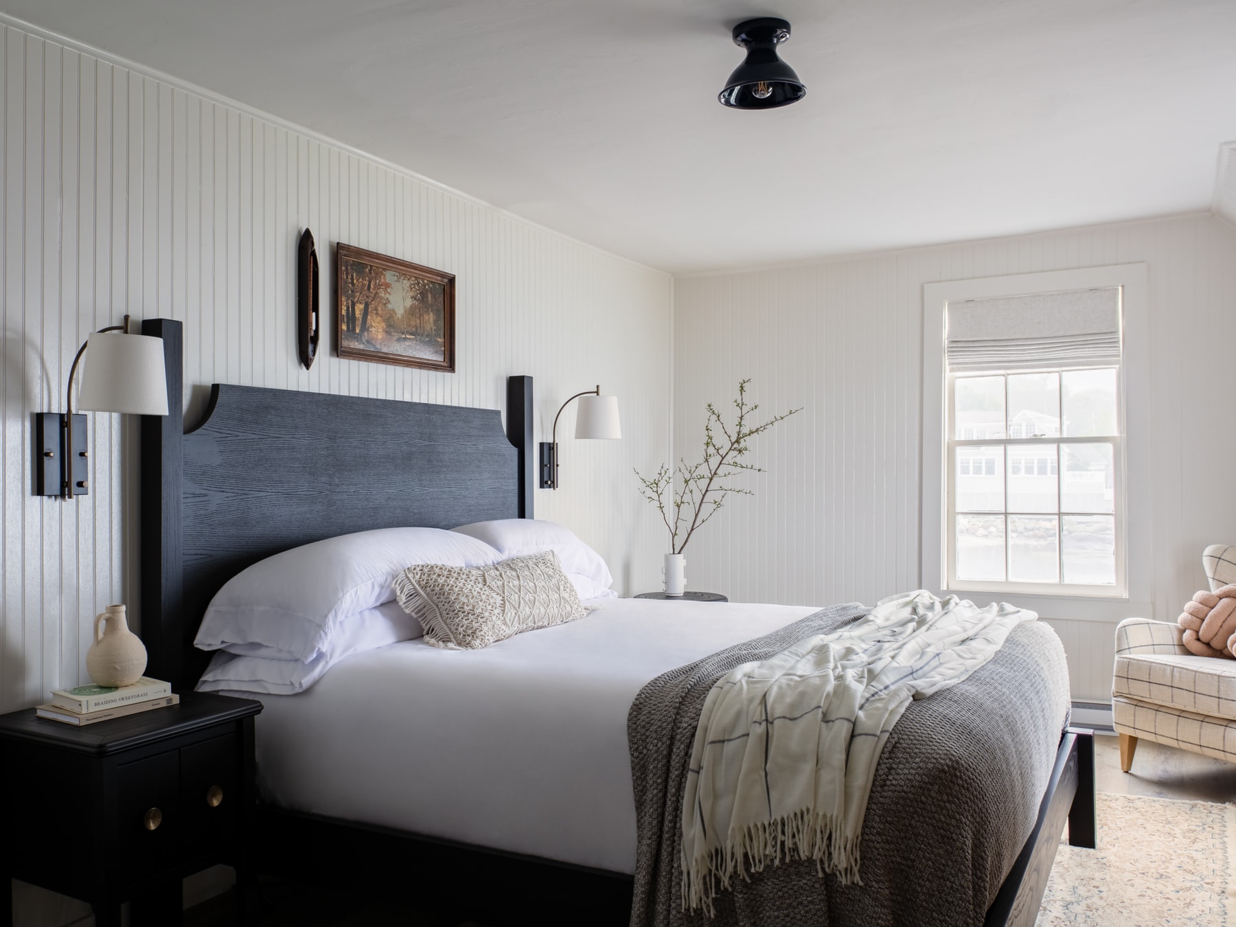 A neatly made bed with white linens and a gray headboard in a room with white walls and a window.