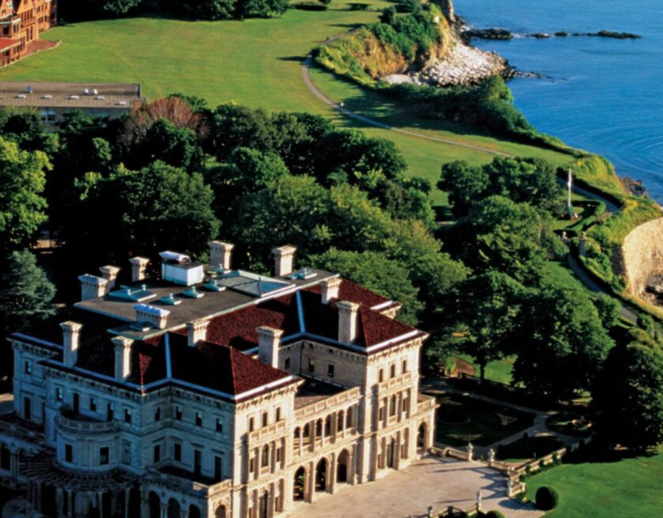 A large building with a red roof and white walls sits on a hill overlooking the ocean.