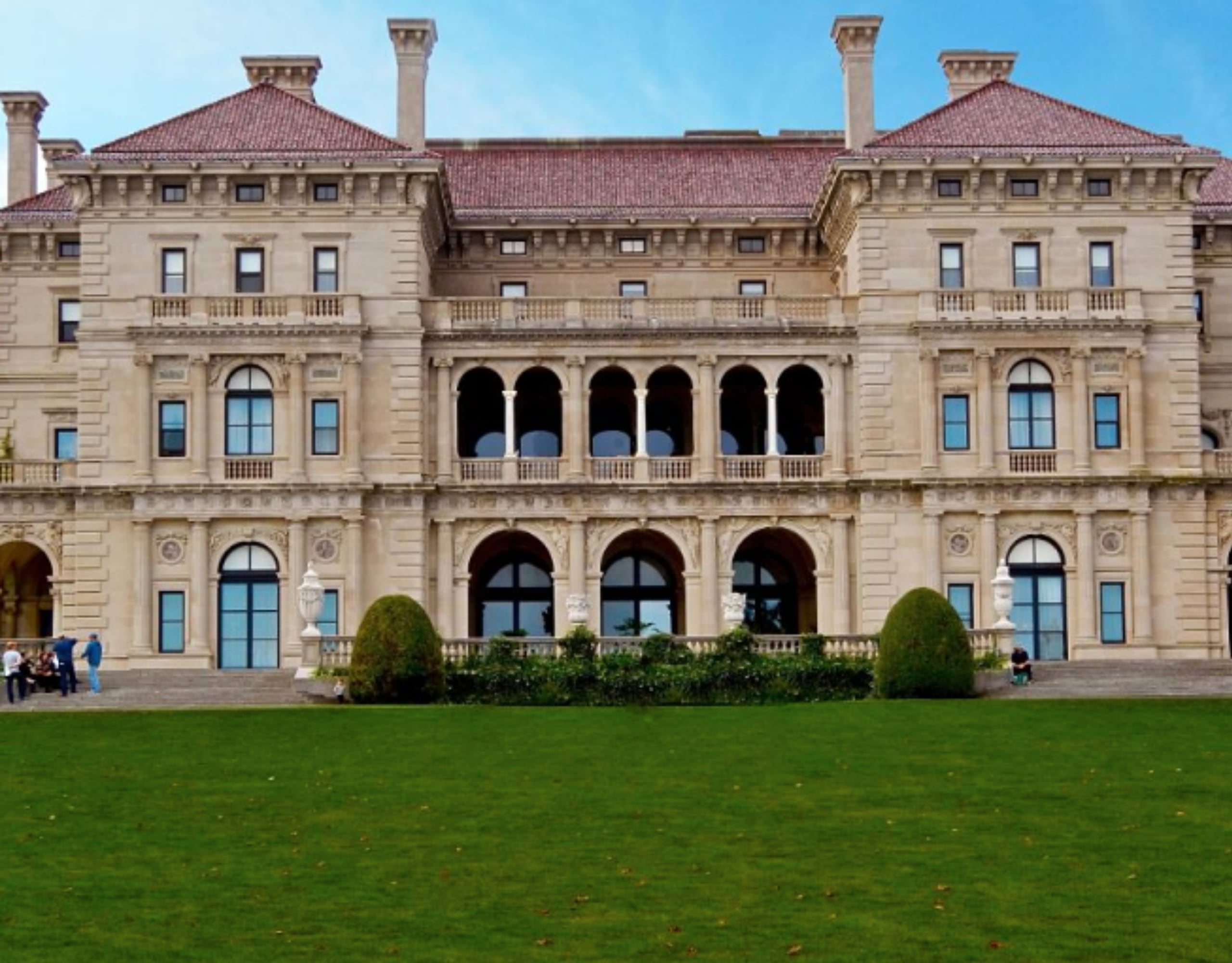 A large, tan-colored building with red-tiled roofs and arched windows and doorways, set against a backdrop of green lawn and blue sky.
