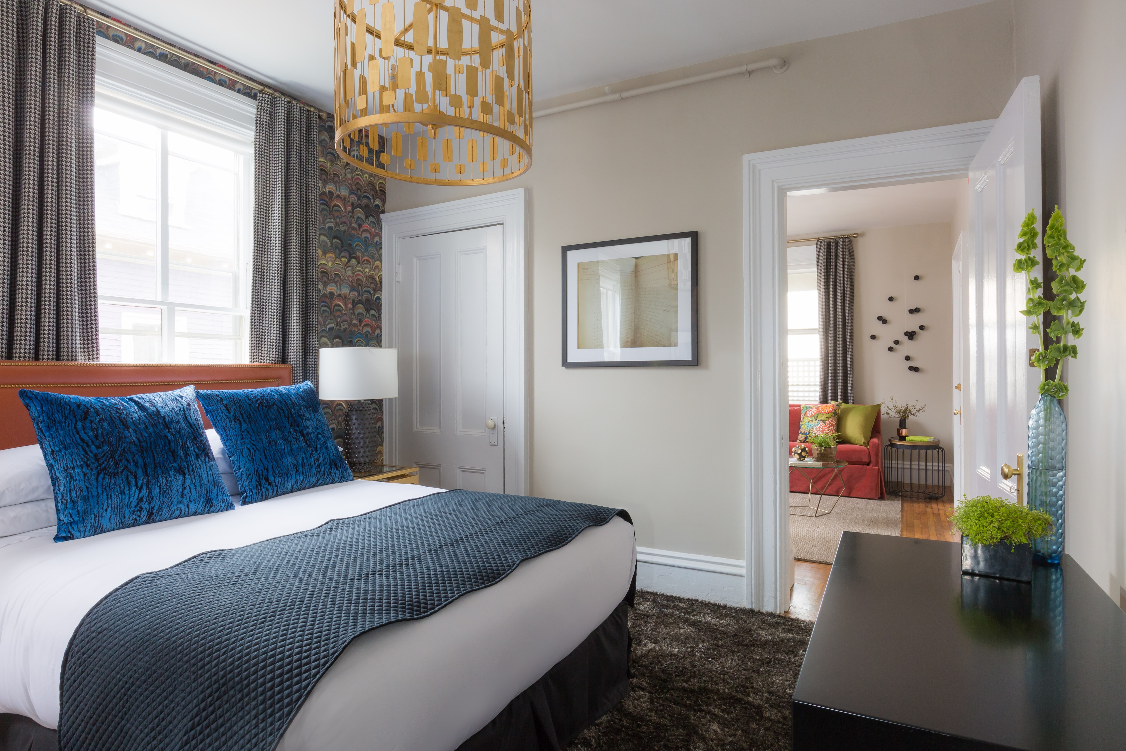A guest room with a bed, window, doorway, chandelier, and plants.