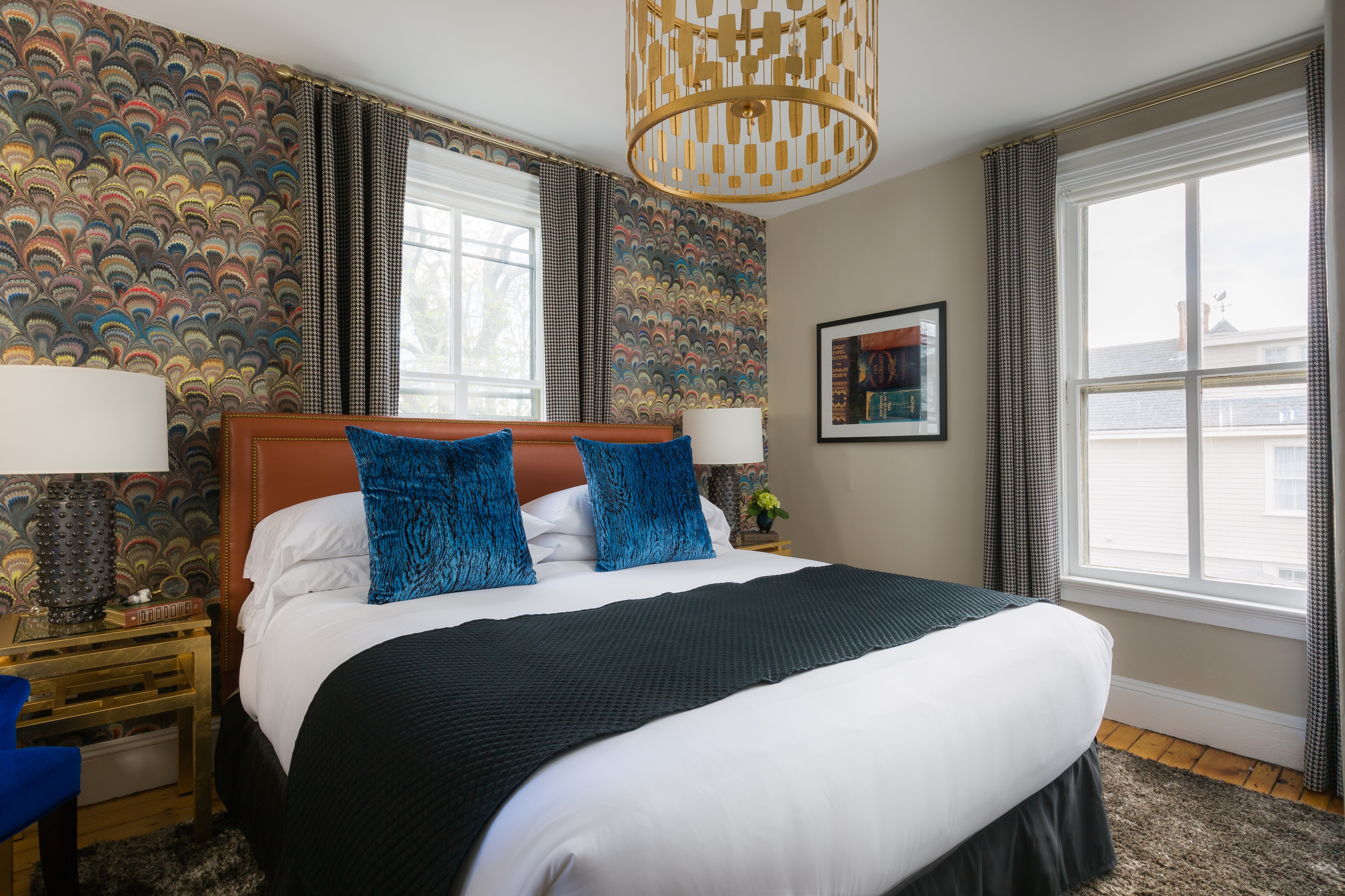 A neatly made bed with blue pillows and a dark throw blanket in a room with colorful wallpaper and two windows.