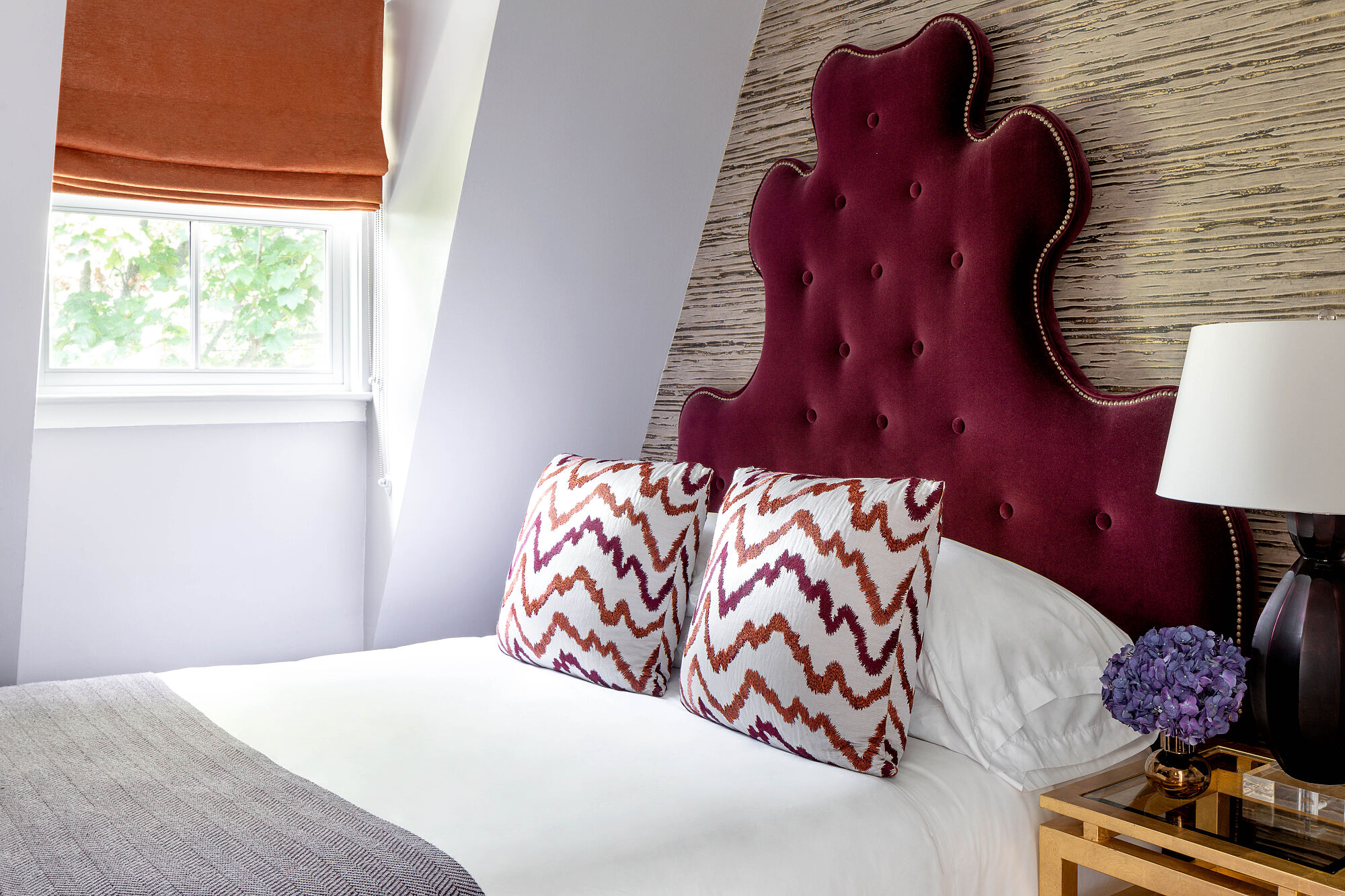 A bed with a tufted headboard, chevron pillows, and white bedding is shown with a lamp and flowers on a nightstand.