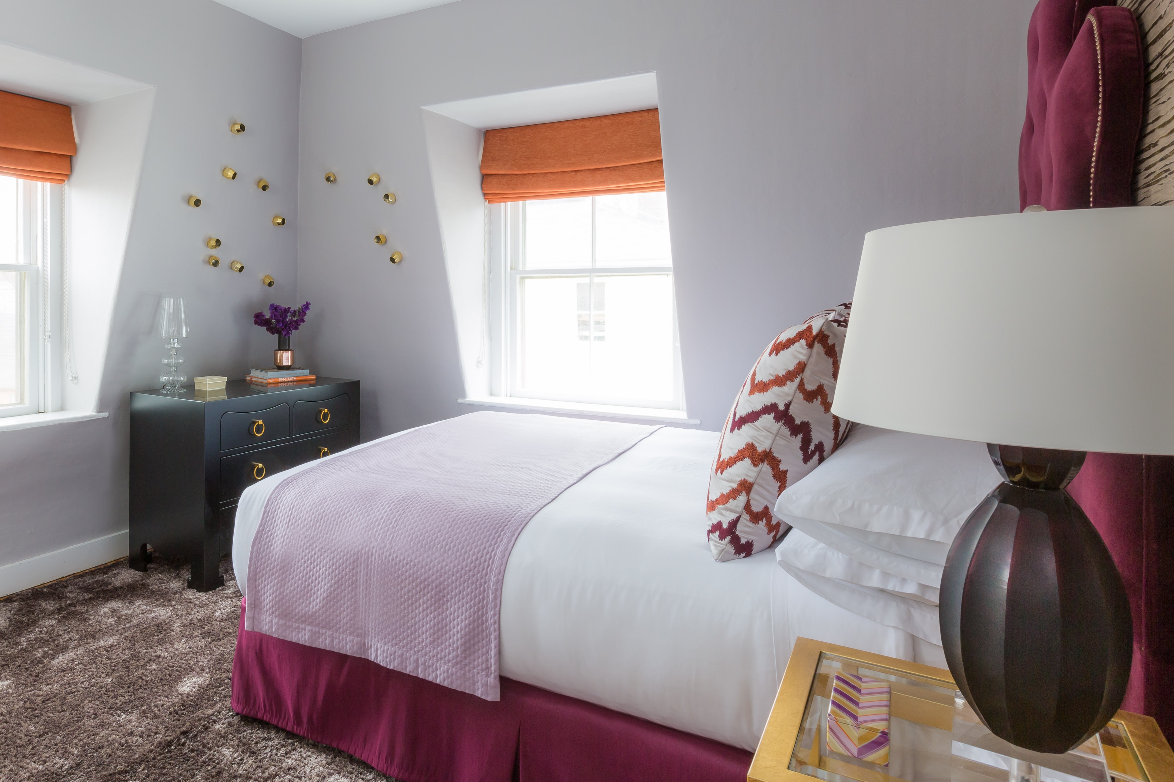 A neatly made bed with white sheets and a pink blanket, a nightstand with a lamp and books, and a window with orange blinds.