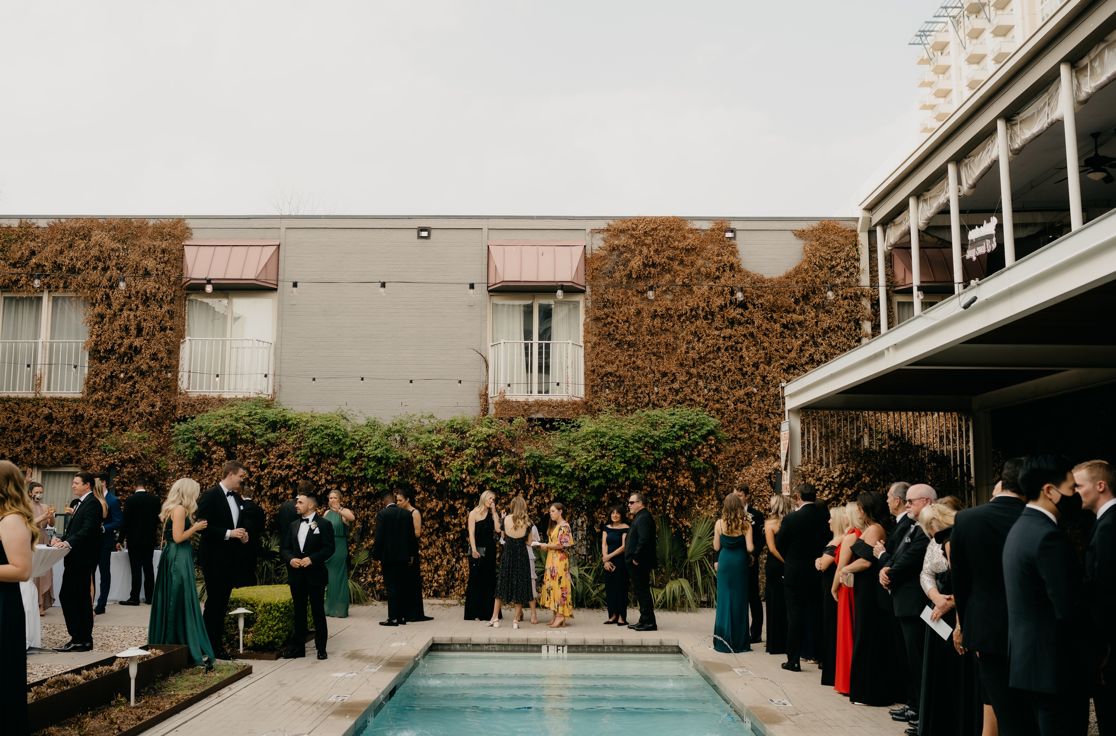 A group of formally dressed people gathered around a pool in an outdoor setting with an ivy-covered wall and building in the background.