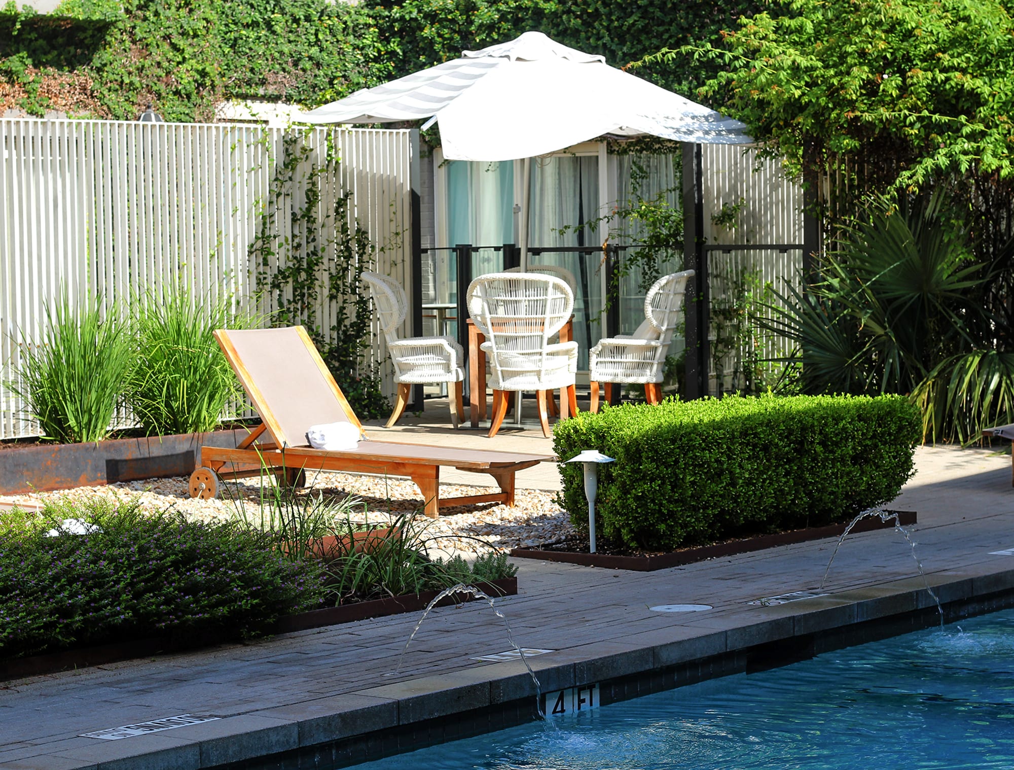 A serene poolside area with a lounge chair, table, and umbrella surrounded by lush greenery and a small fountain.