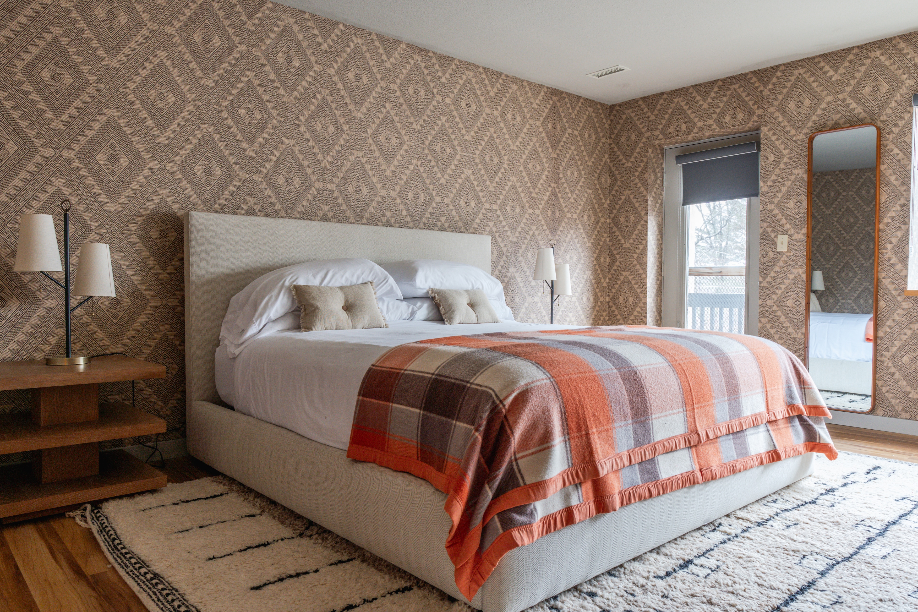 A neatly made bed with white linens and an orange plaid blanket, with a nightstand and mirror in a room with patterned wallpaper.