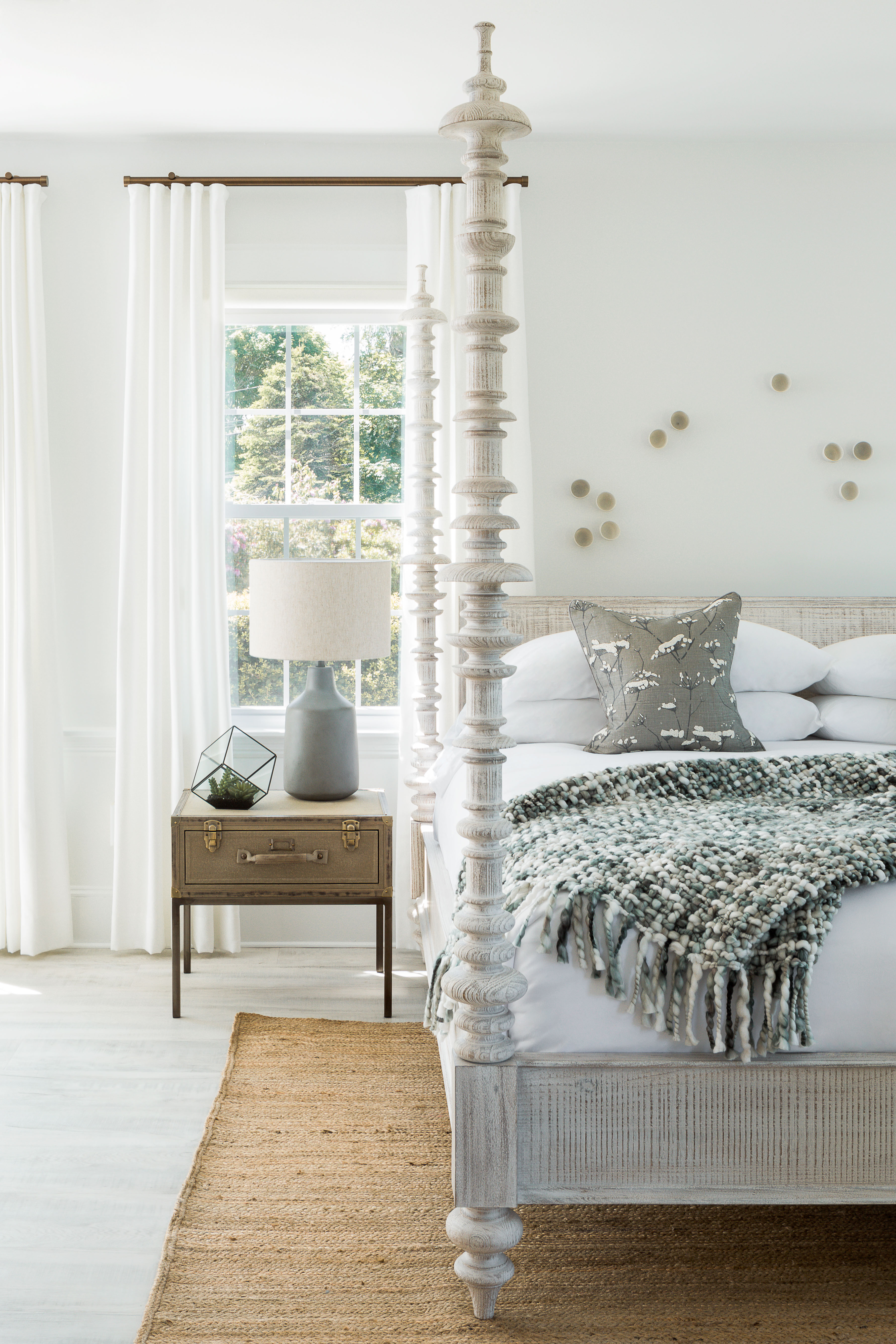 A white four-poster bed with a throw blanket and pillows, a nightstand with a lamp, and a window with curtains.