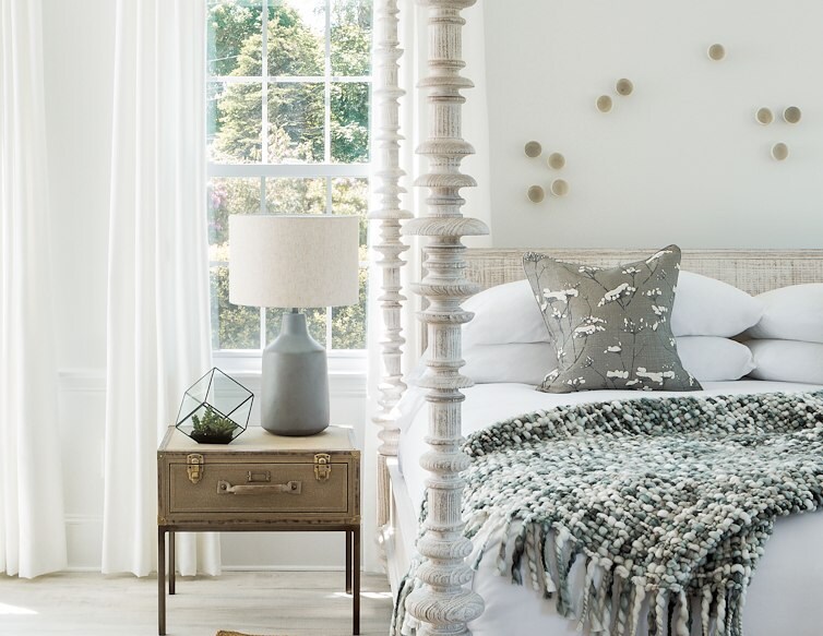 Bright guest room with a carved four-poster bed, white curtains, textured bedding, and a bedside lamp beside a sunny window.