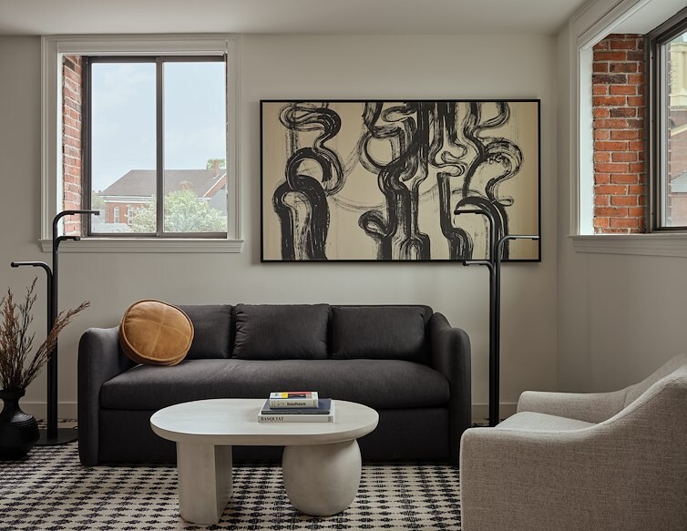 Modern hotel lounge with a dark sofa, sculptural coffee table, abstract artwork, and large windows framed by exposed brick.