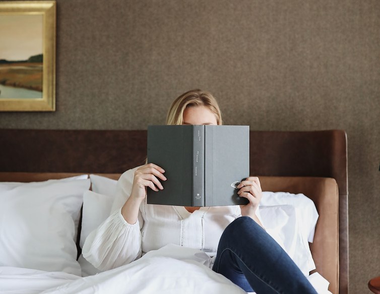Guest relaxing in bed while reading, with breakfast, coffee, and a croissant served on a tray in the foreground.