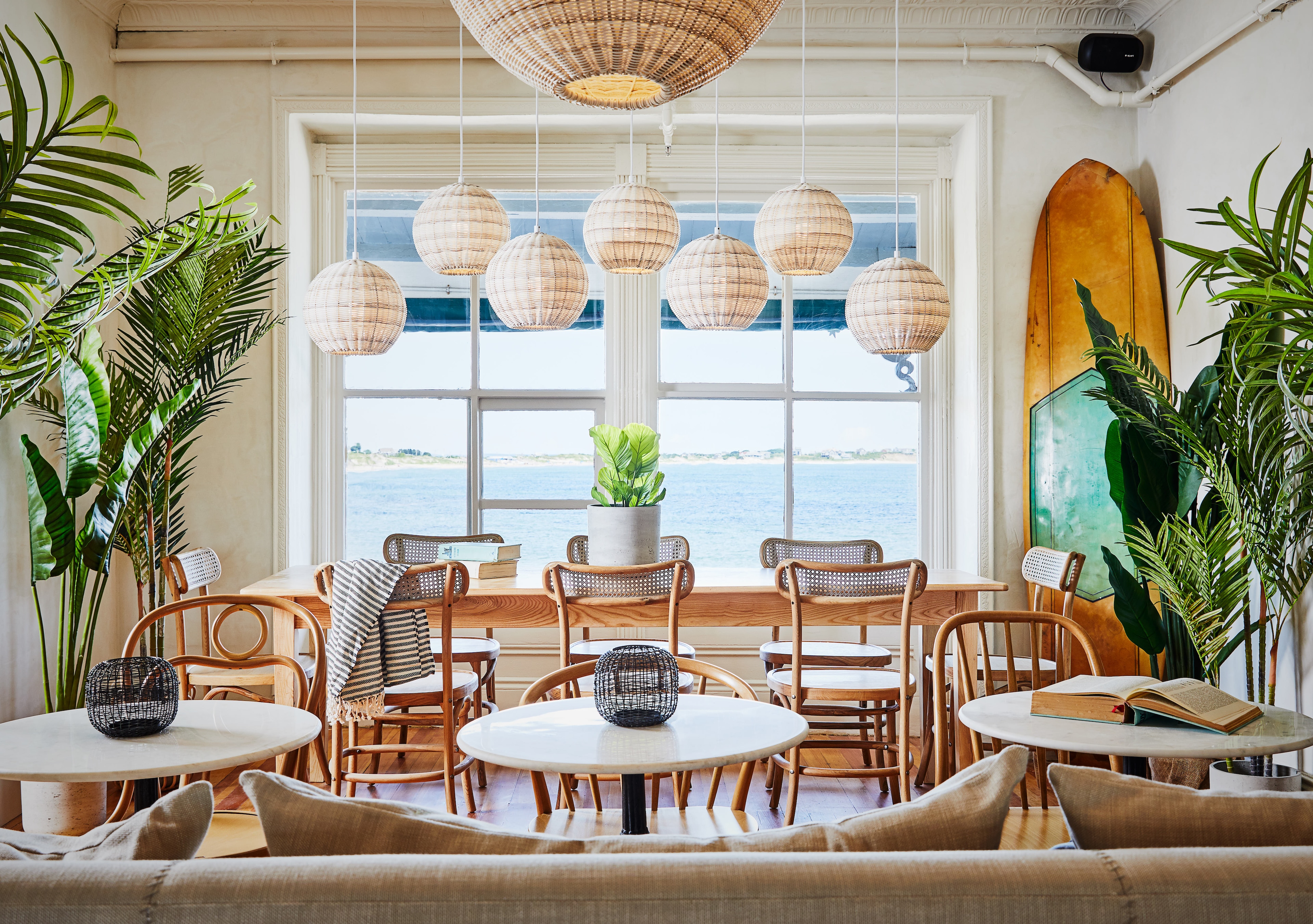 A bright and airy lounge area with round tables, wooden chairs, and wicker light fixtures, overlooking the water.
