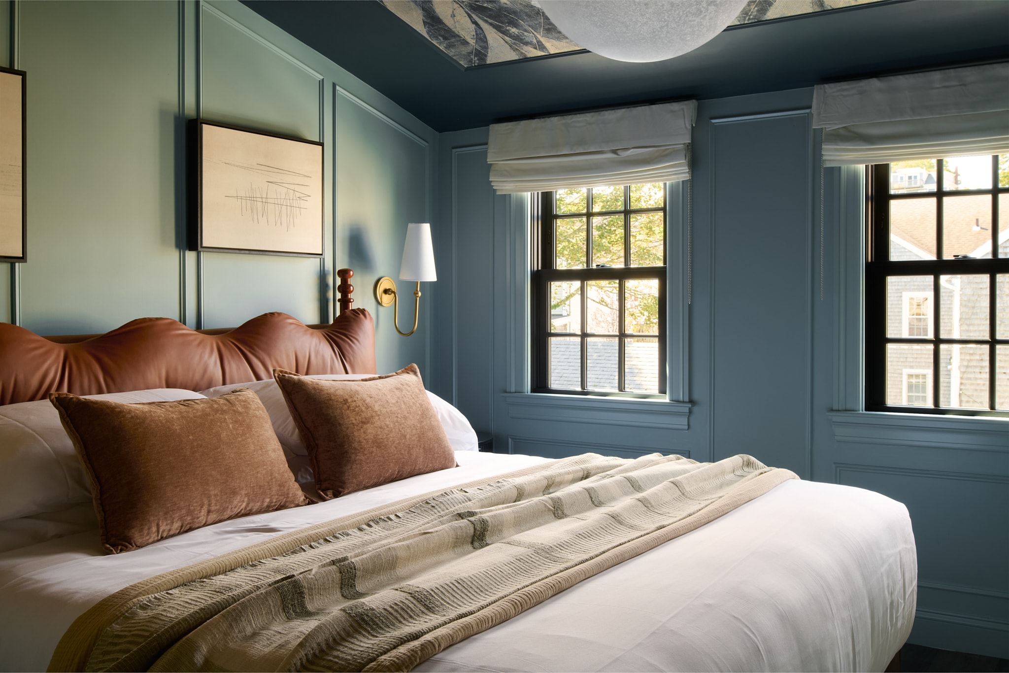 A neatly made bed with brown pillows and a white comforter in a room with blue walls and two windows.