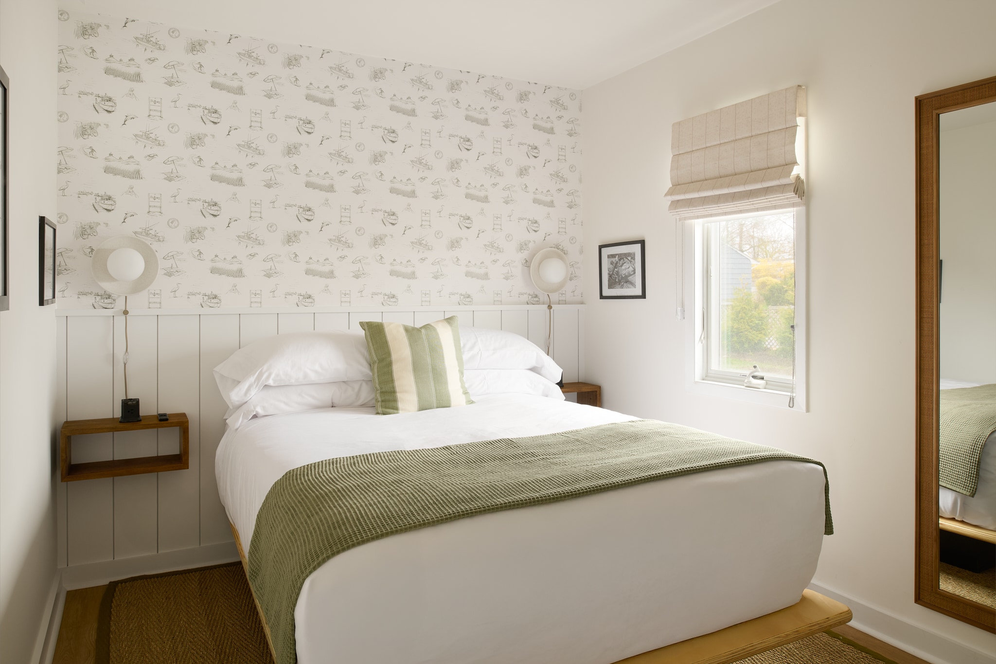 A neatly made bed with white sheets and green accents in a room with patterned wallpaper and a window.