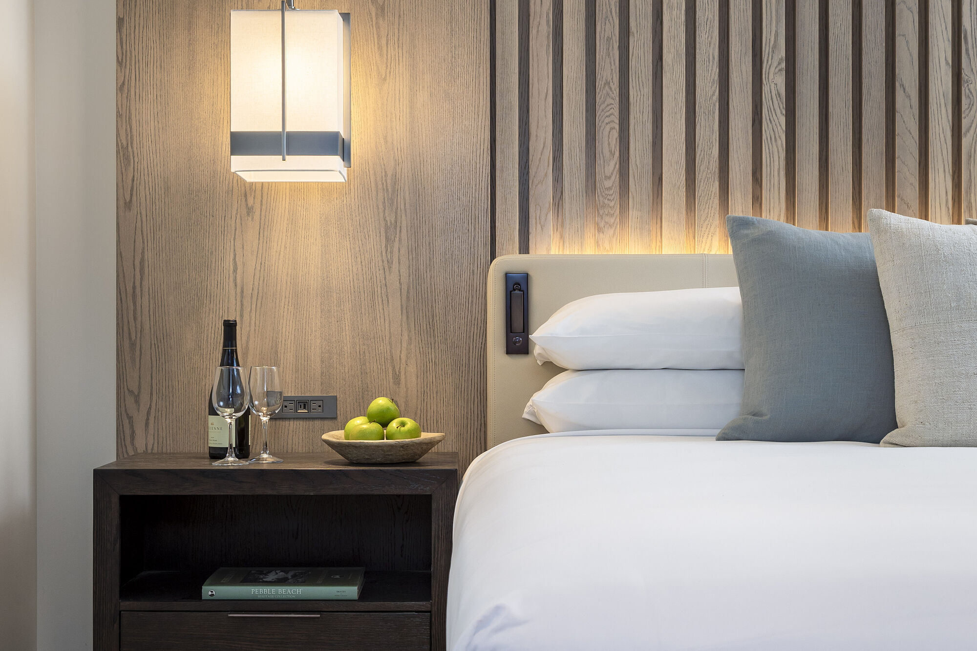 A close-up of a bed with a nightstand, lamp, book, wine, and apples.