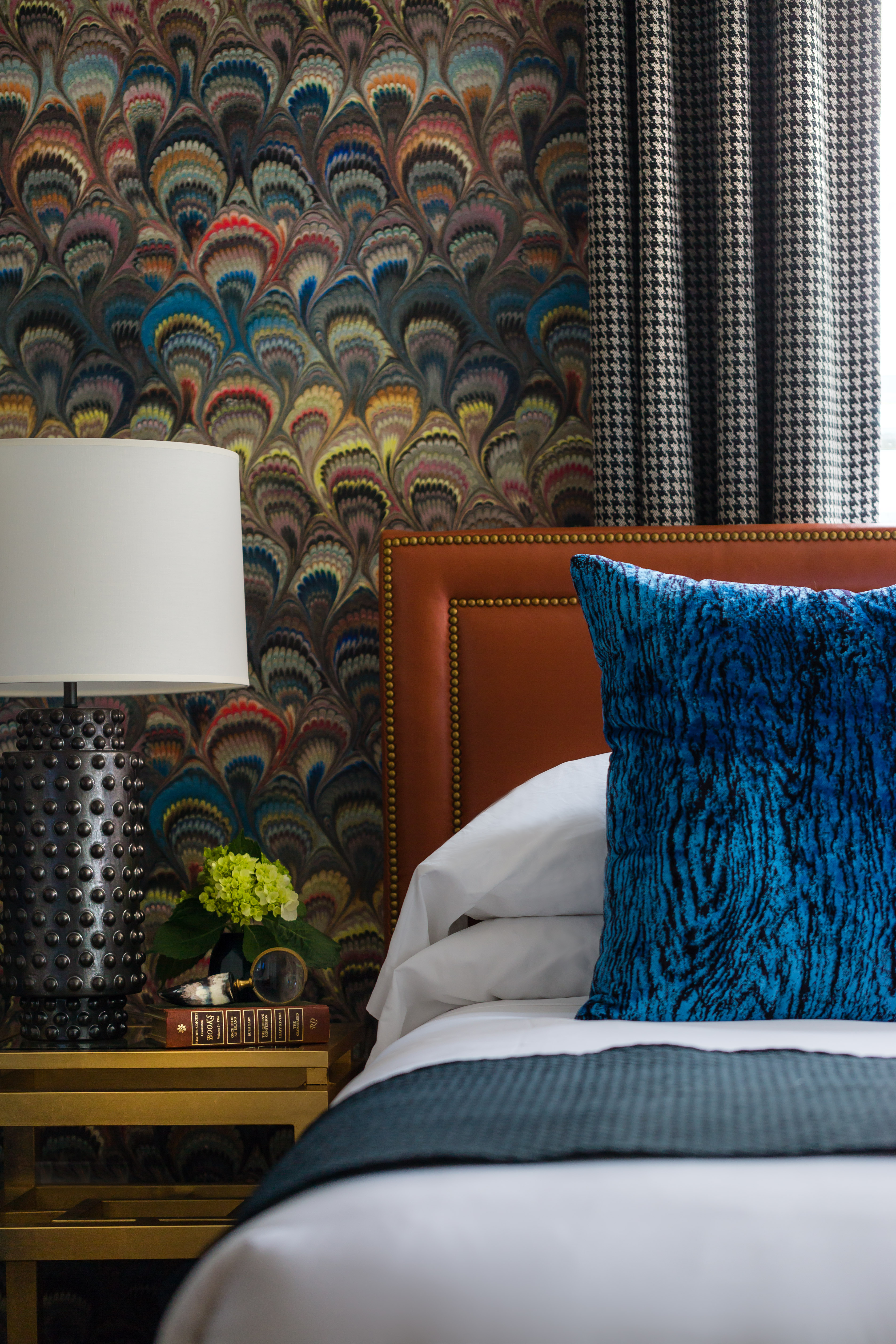 A close-up of a bed with a blue pillow and a brown headboard in front of a floral wallpaper.