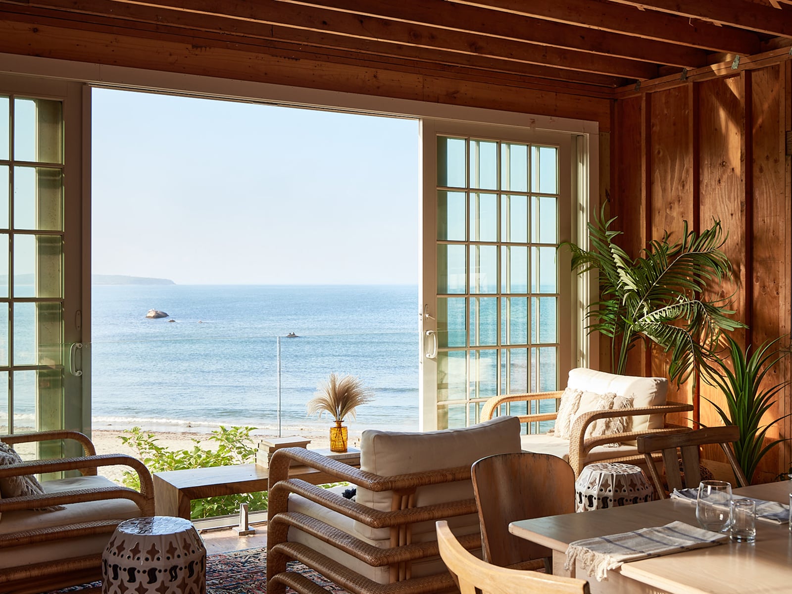 A beachside dining area with wood paneling, large windows, and wicker furniture overlooking the ocean.