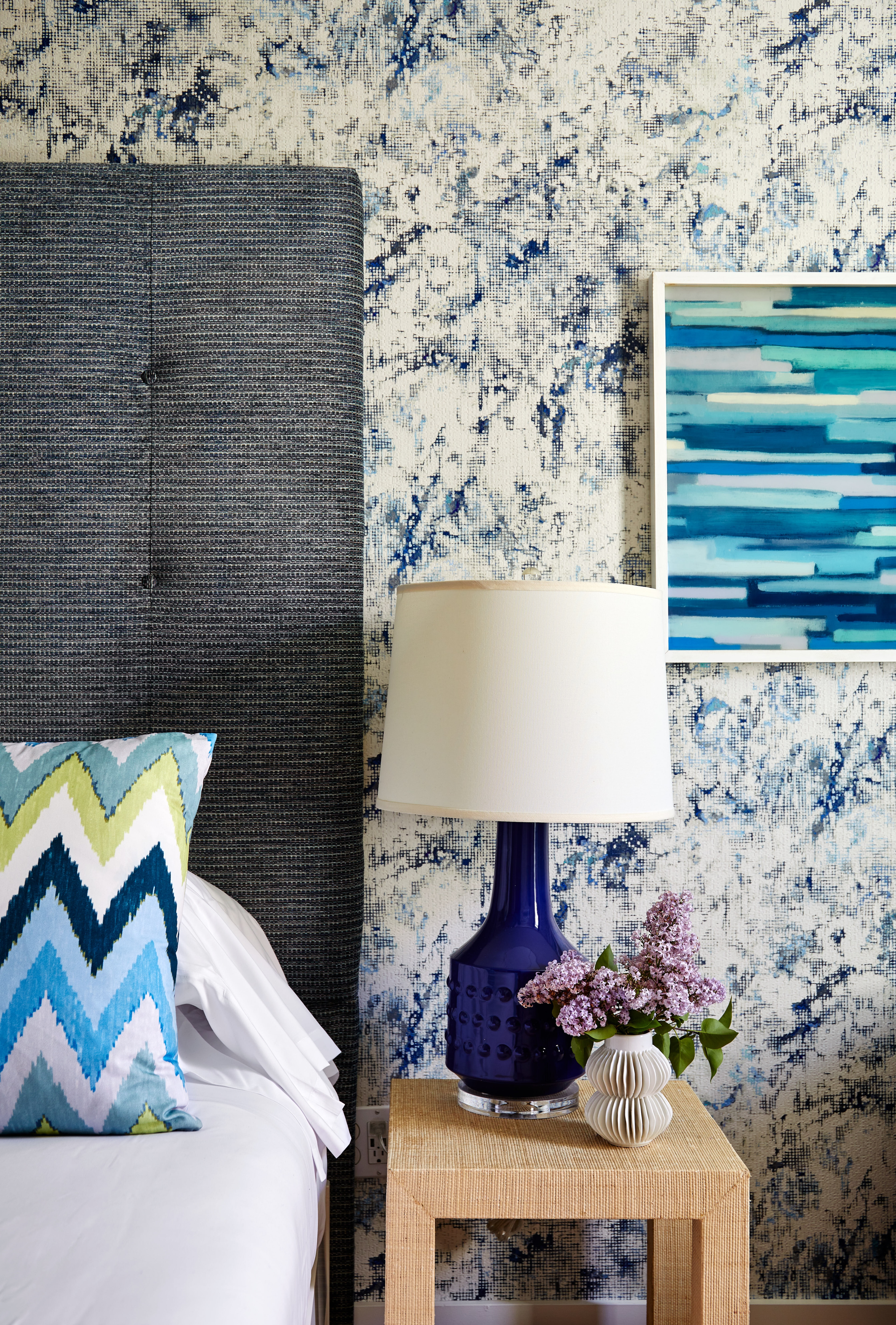 A bed with a white comforter and blue and green pillow, a nightstand with a blue lamp and flowers, and blue and white wallpaper.