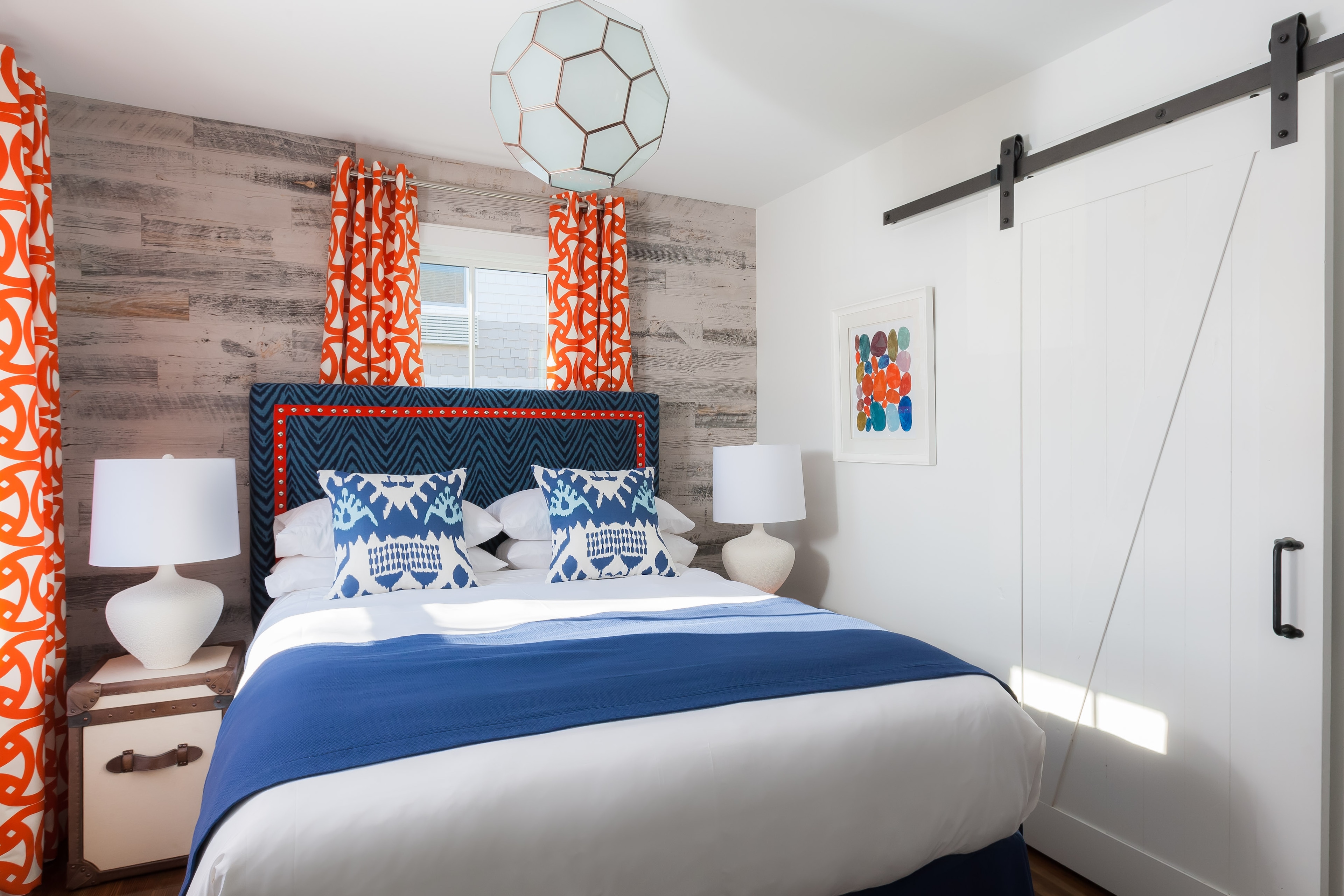 A neatly made bed with blue and white bedding and pillows, with a wooden headboard and a window with orange curtains.
