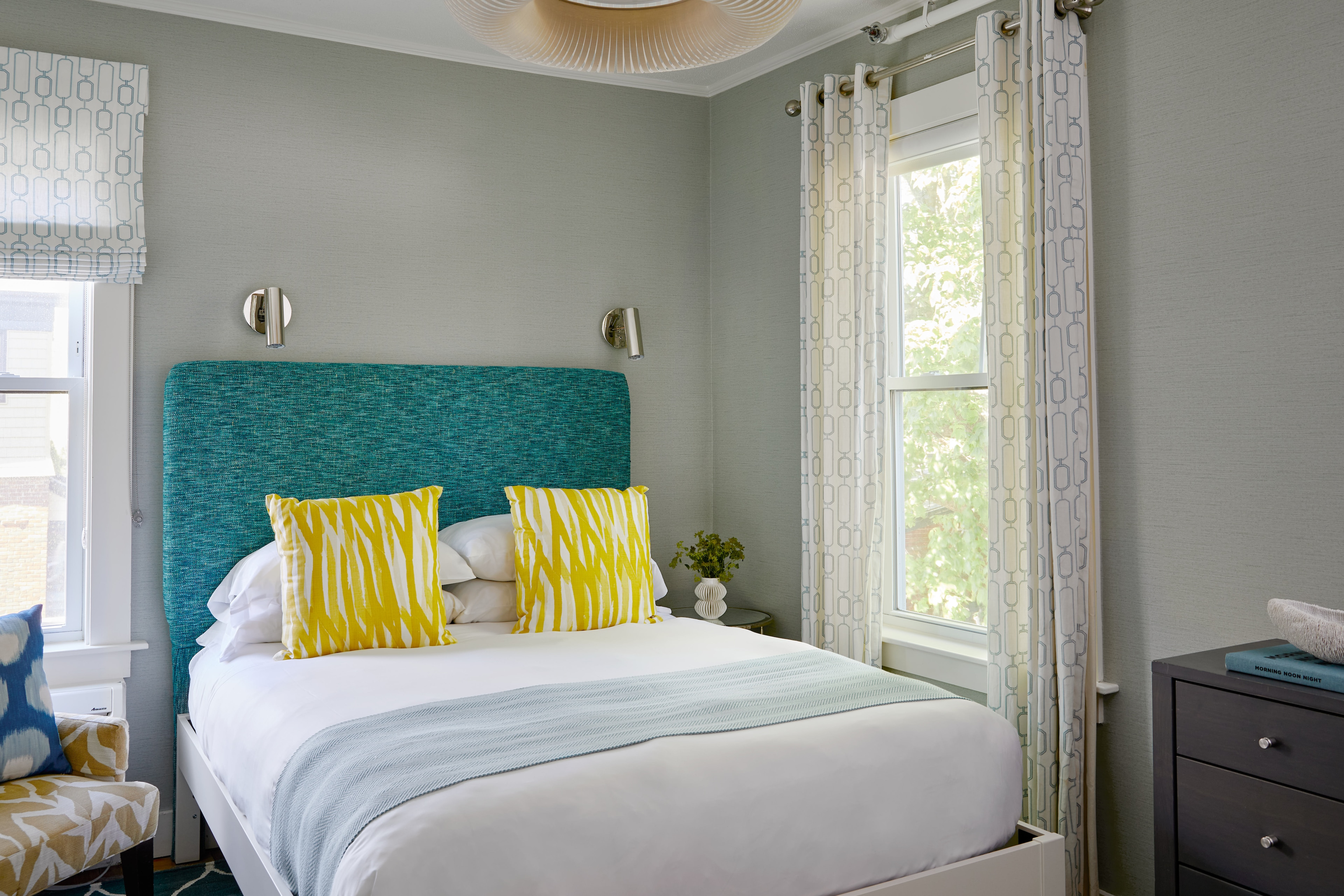 A neatly made bed with white sheets and yellow pillows in a room with gray walls and two windows.
