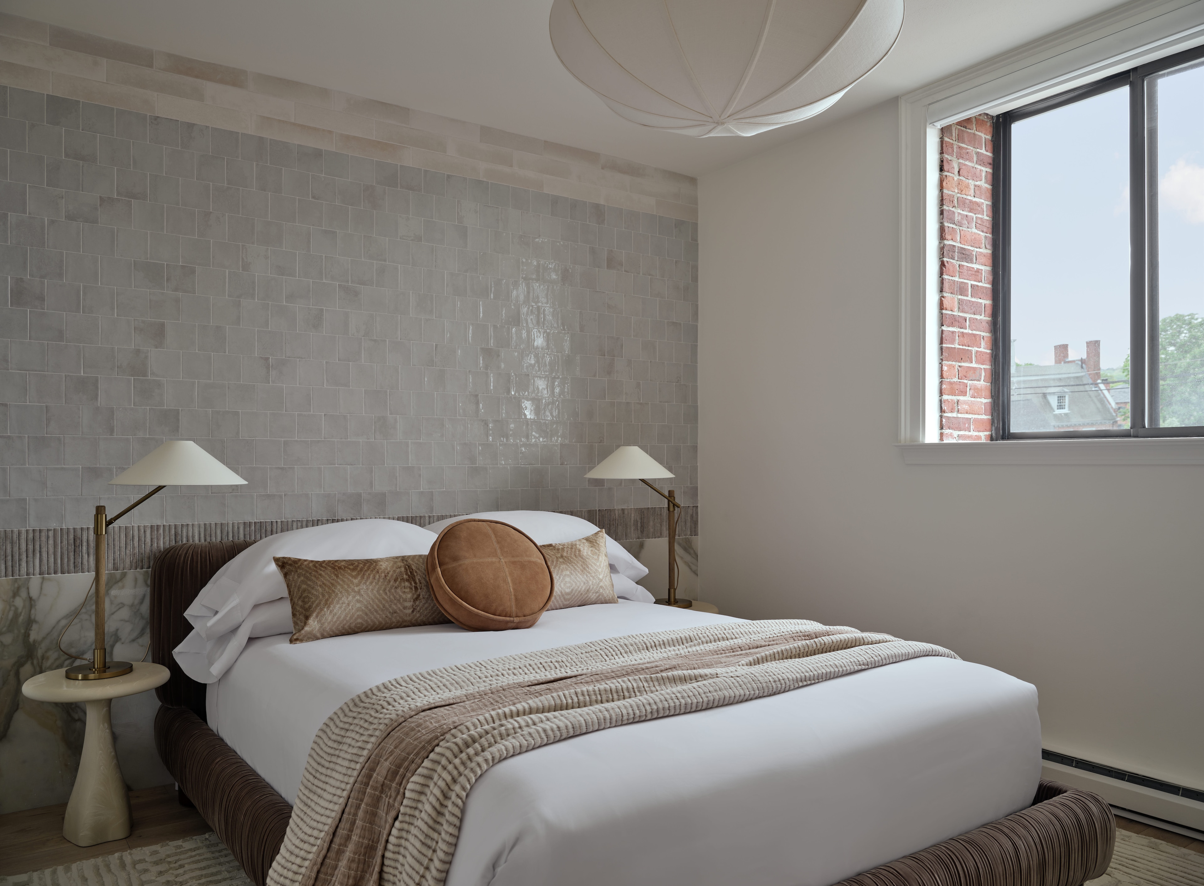 A neatly made bed with pillows and a throw blanket in a room with a tiled accent wall and a window.