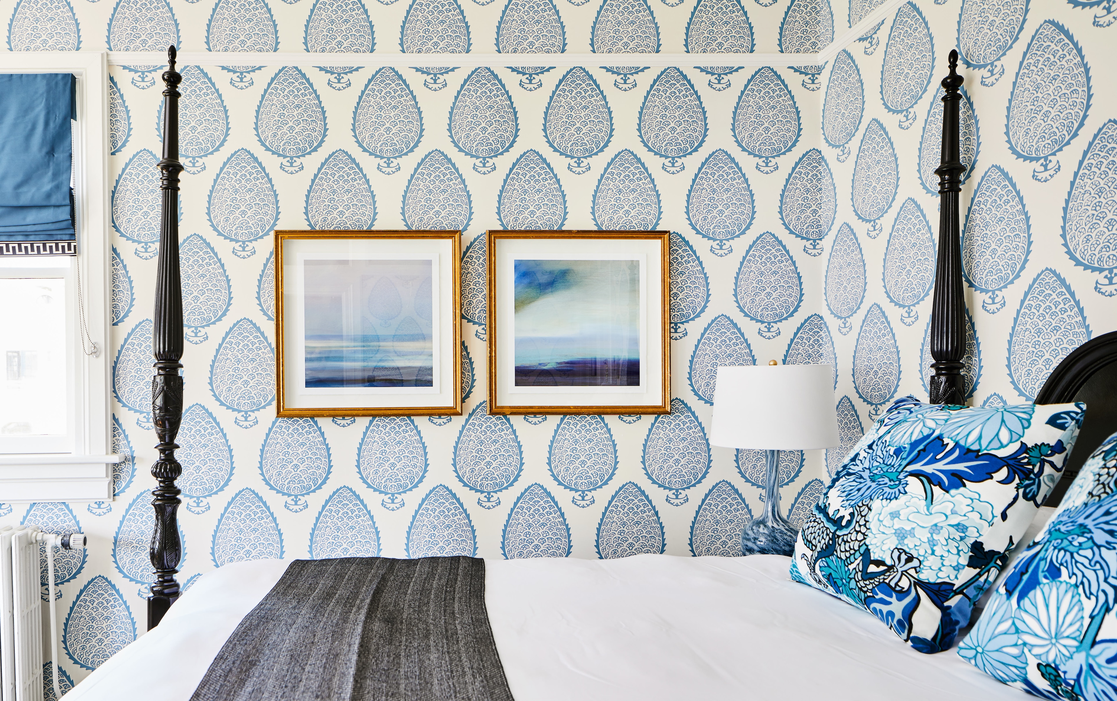 A bed with blue and white pillows and a blue and white wallpaper background with two pieces of framed artwork above the bed.