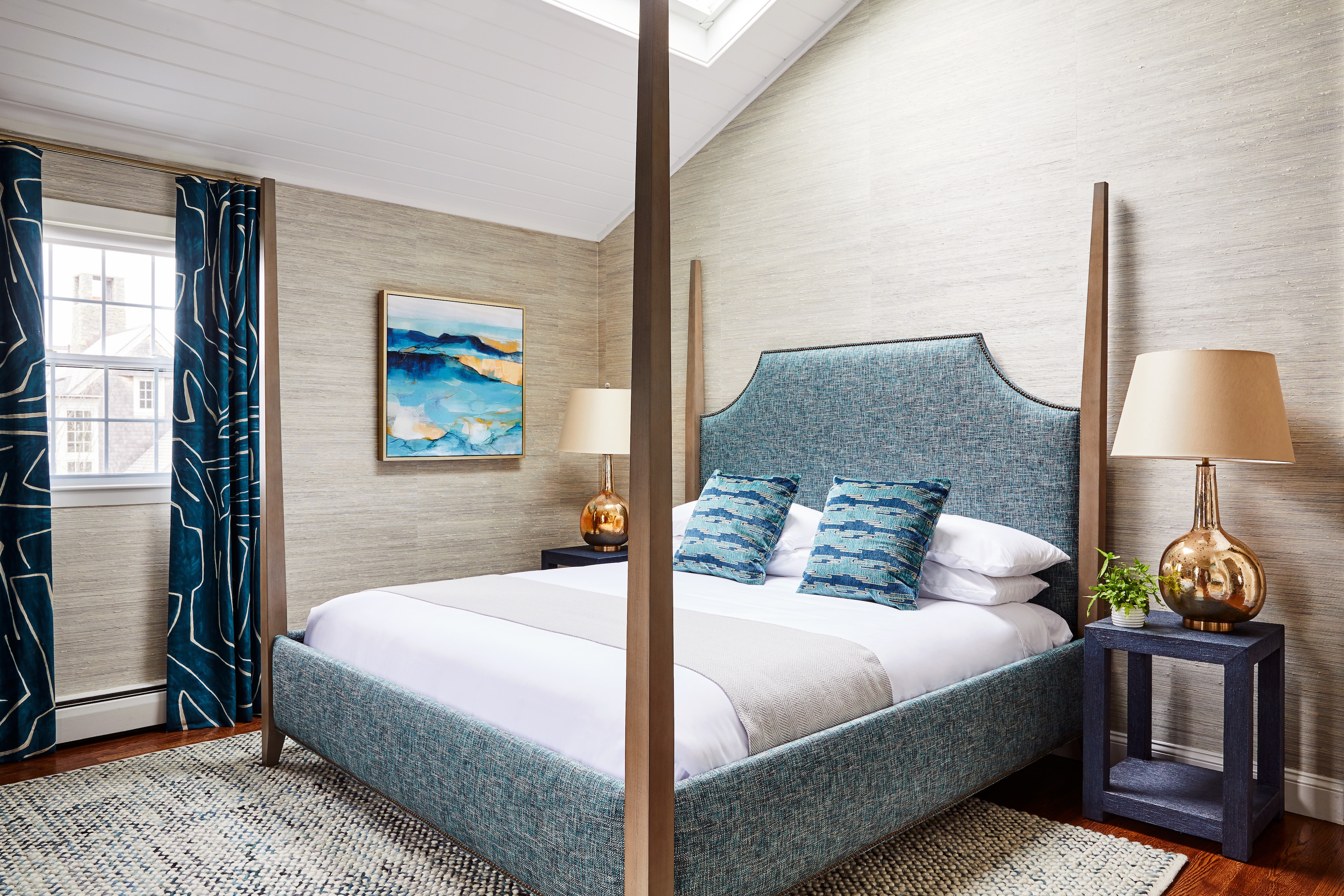 A hotel room with a blue upholstered bed, white bedding, and a painting on the wall.