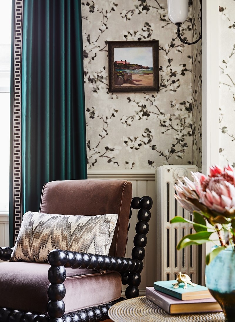 A cozy room with a velvet chair, floral wallpaper, and a vase with flowers.
