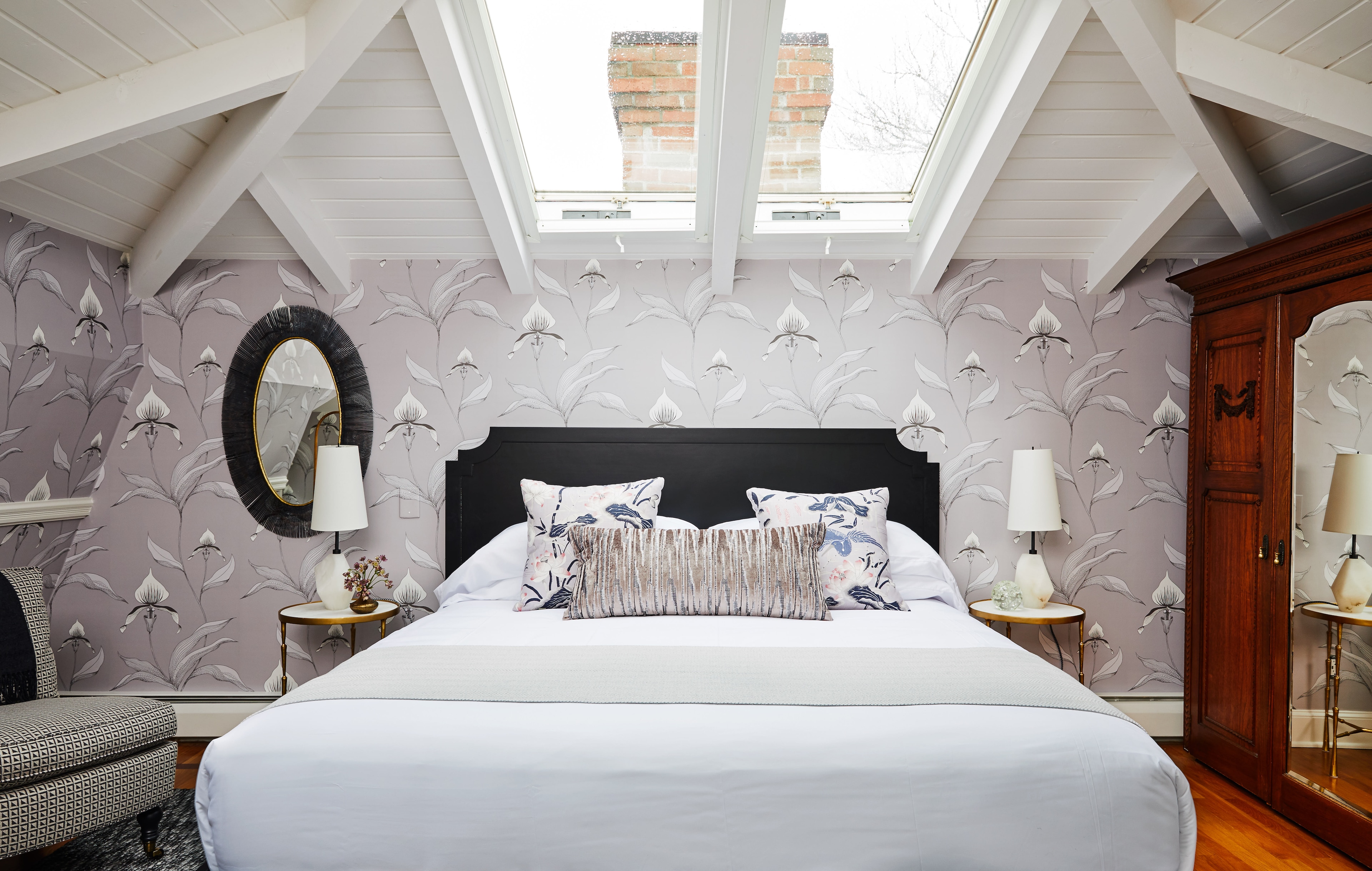 A neatly made bed with pillows and a floral patterned wallpaper in a room with a skylight.