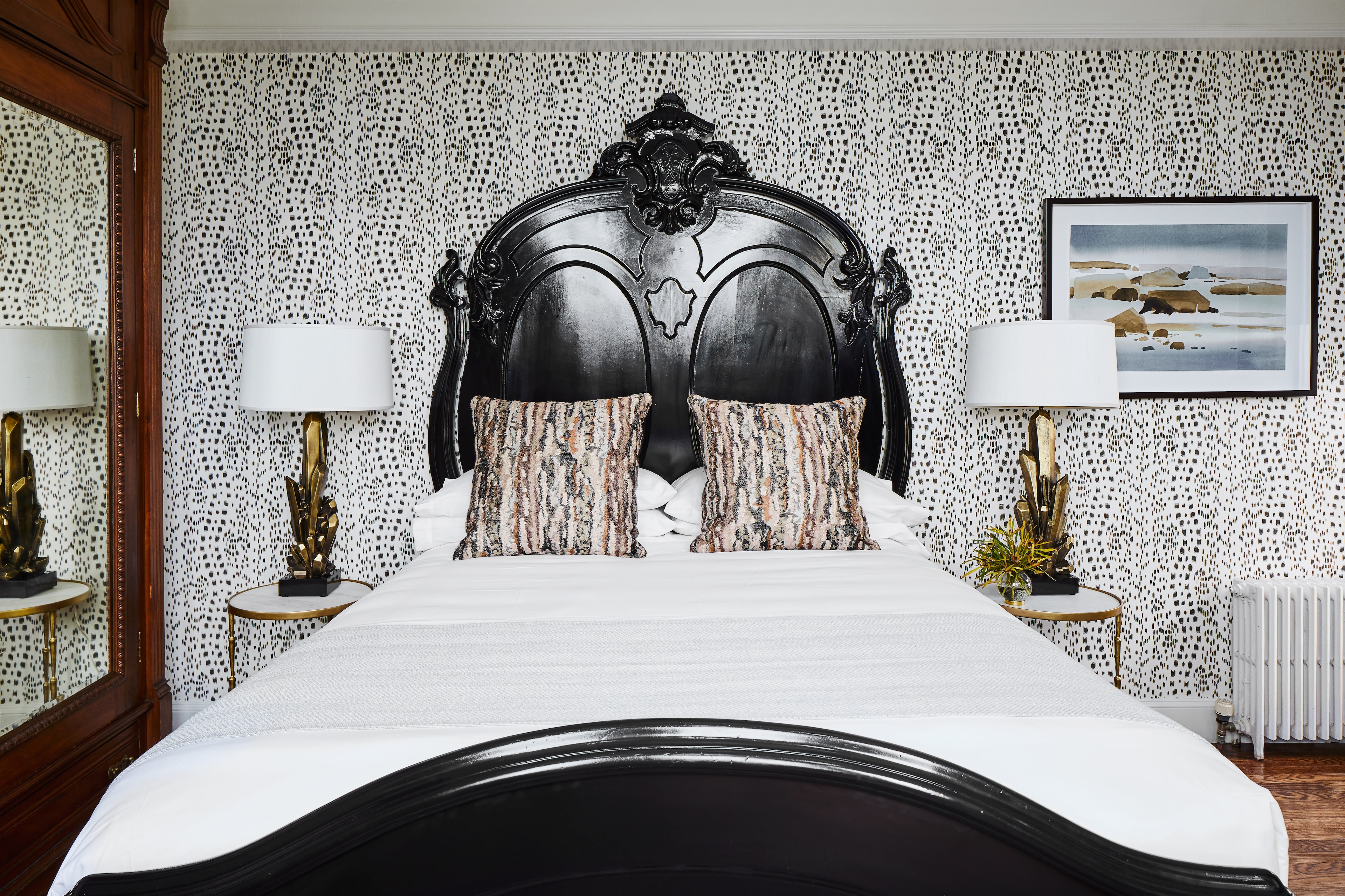 A close-up view of a bed with an ornate headboard, white bedding, and patterned wallpaper.