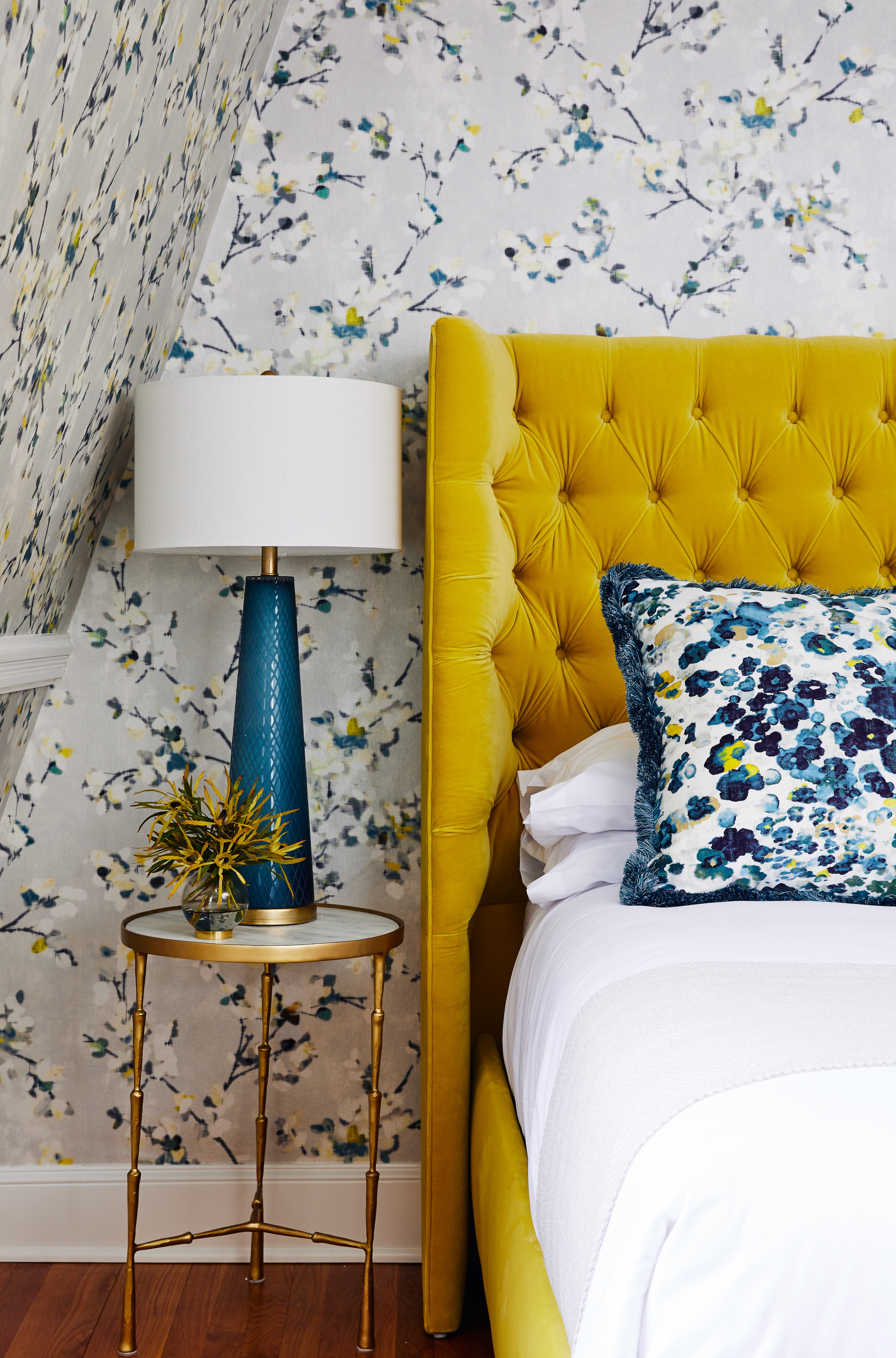 A close-up of a bed with a yellow headboard, white bedding, and a blue lamp on a small table.