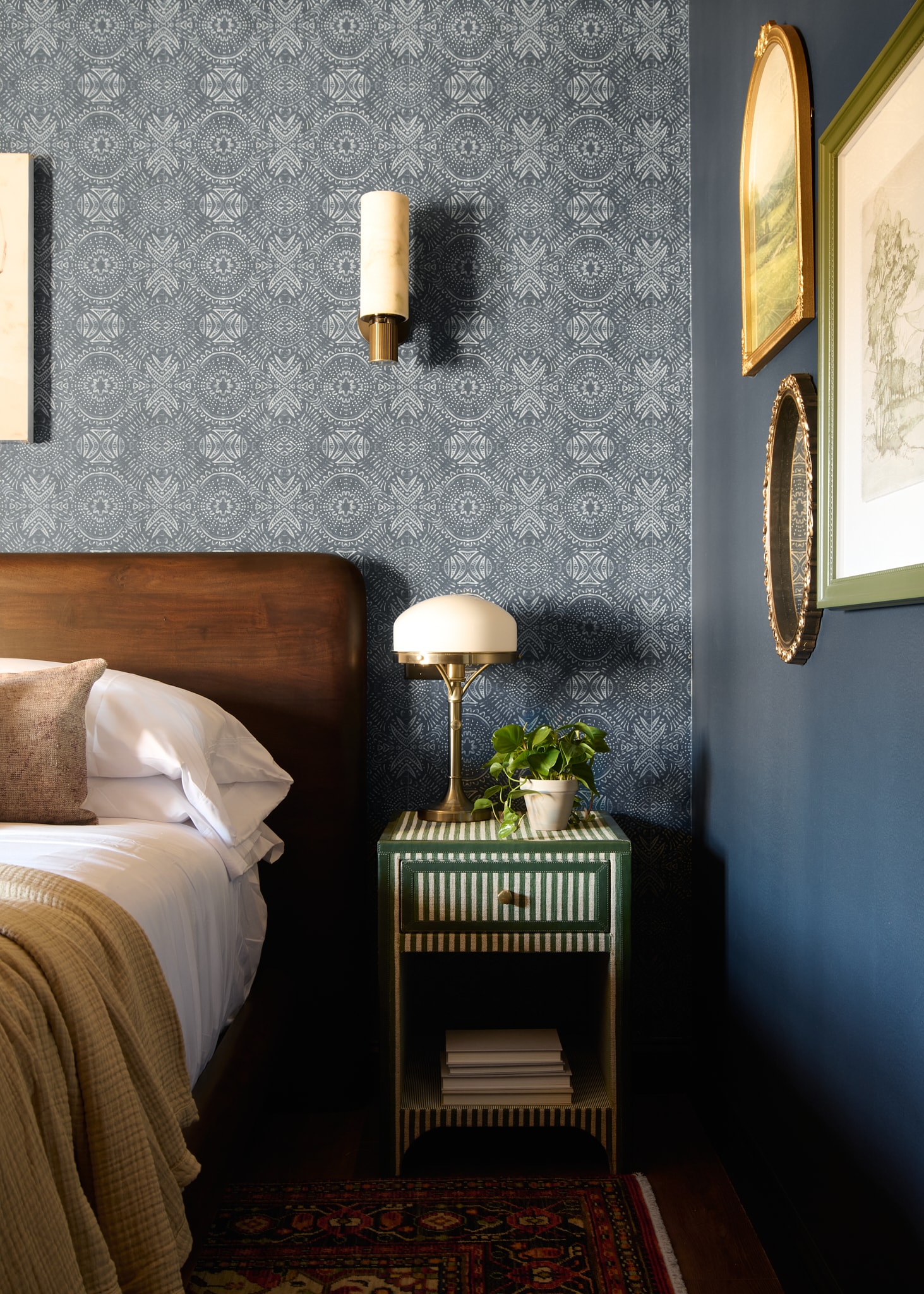 A neatly made bed with a wooden headboard, a green nightstand with a lamp and plant, and blue patterned wallpaper.