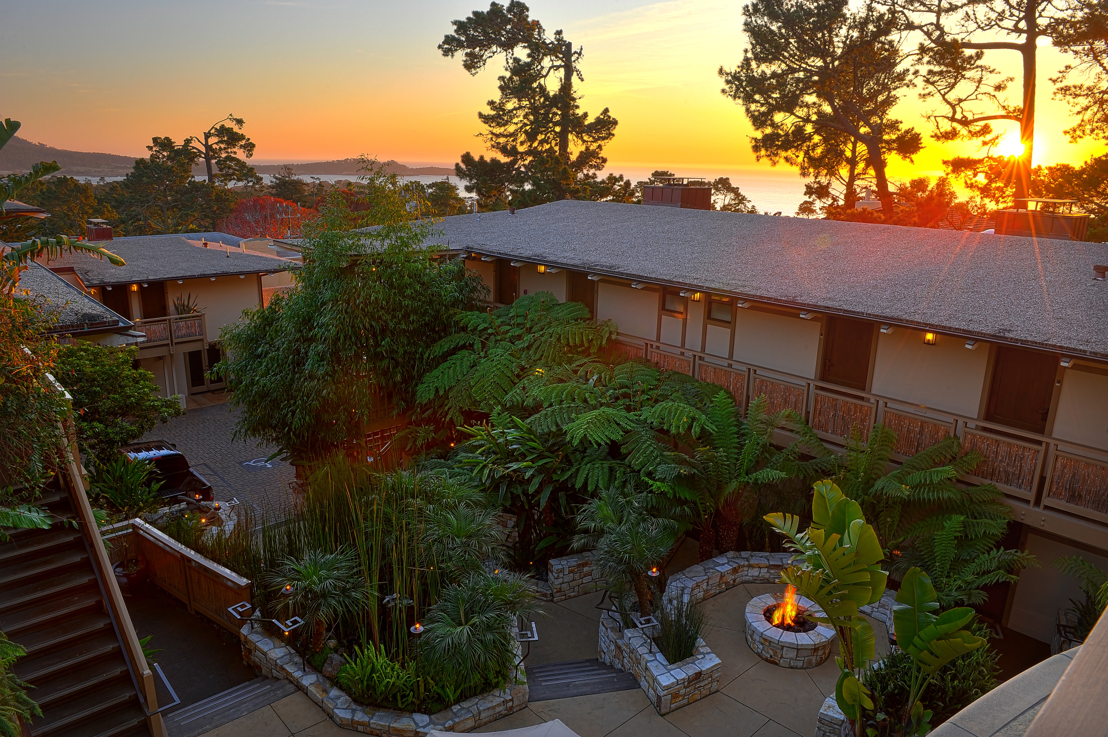 A two-story building with a gray roof surrounded by trees and greenery, with a fire pit in the foreground at sunset.