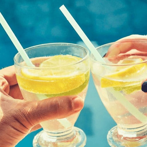 two hands holding glasses with yellow drinks and straws