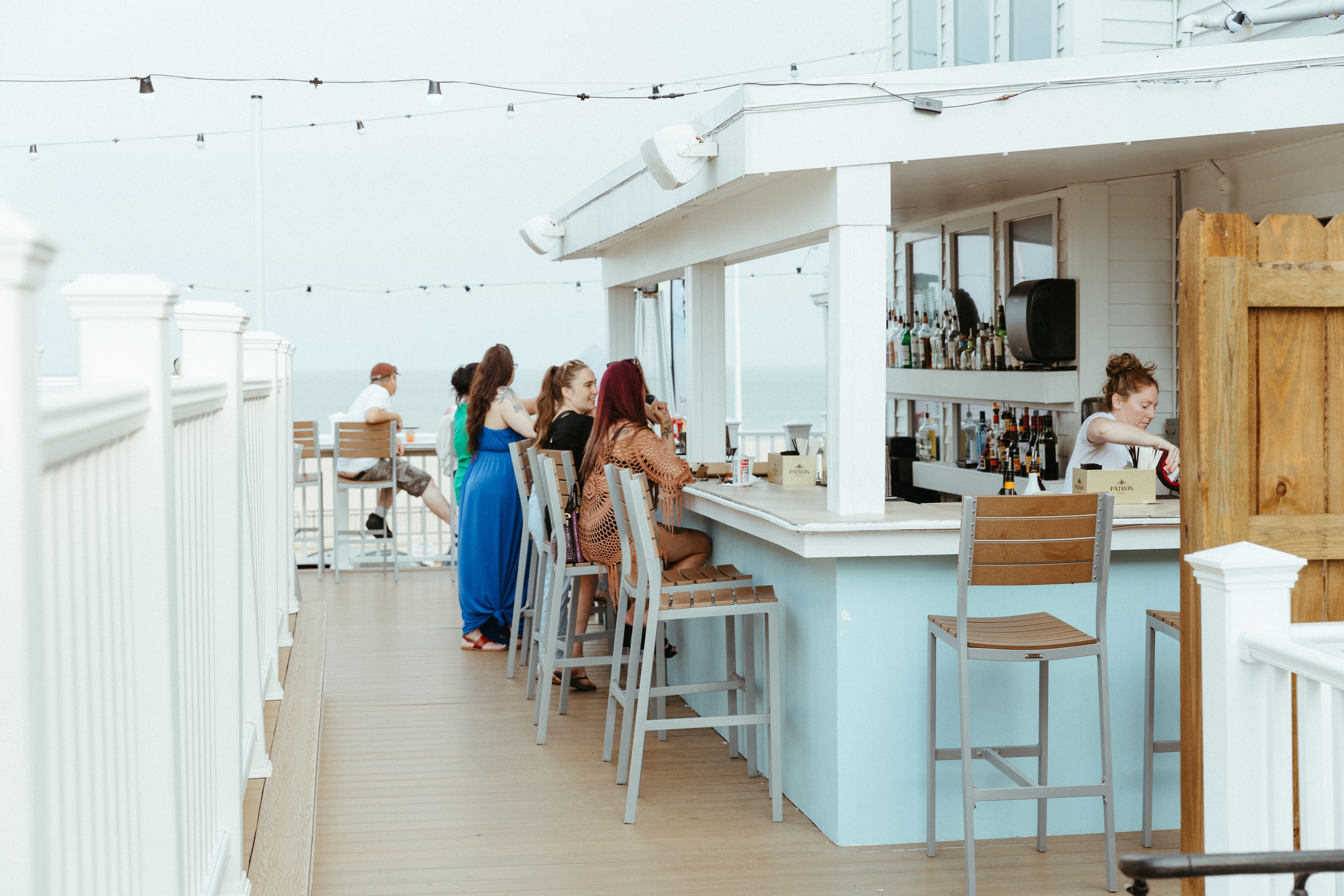 A group of people sitting at an outdoor bar with a light blue base and white countertop, surrounded by string lights.