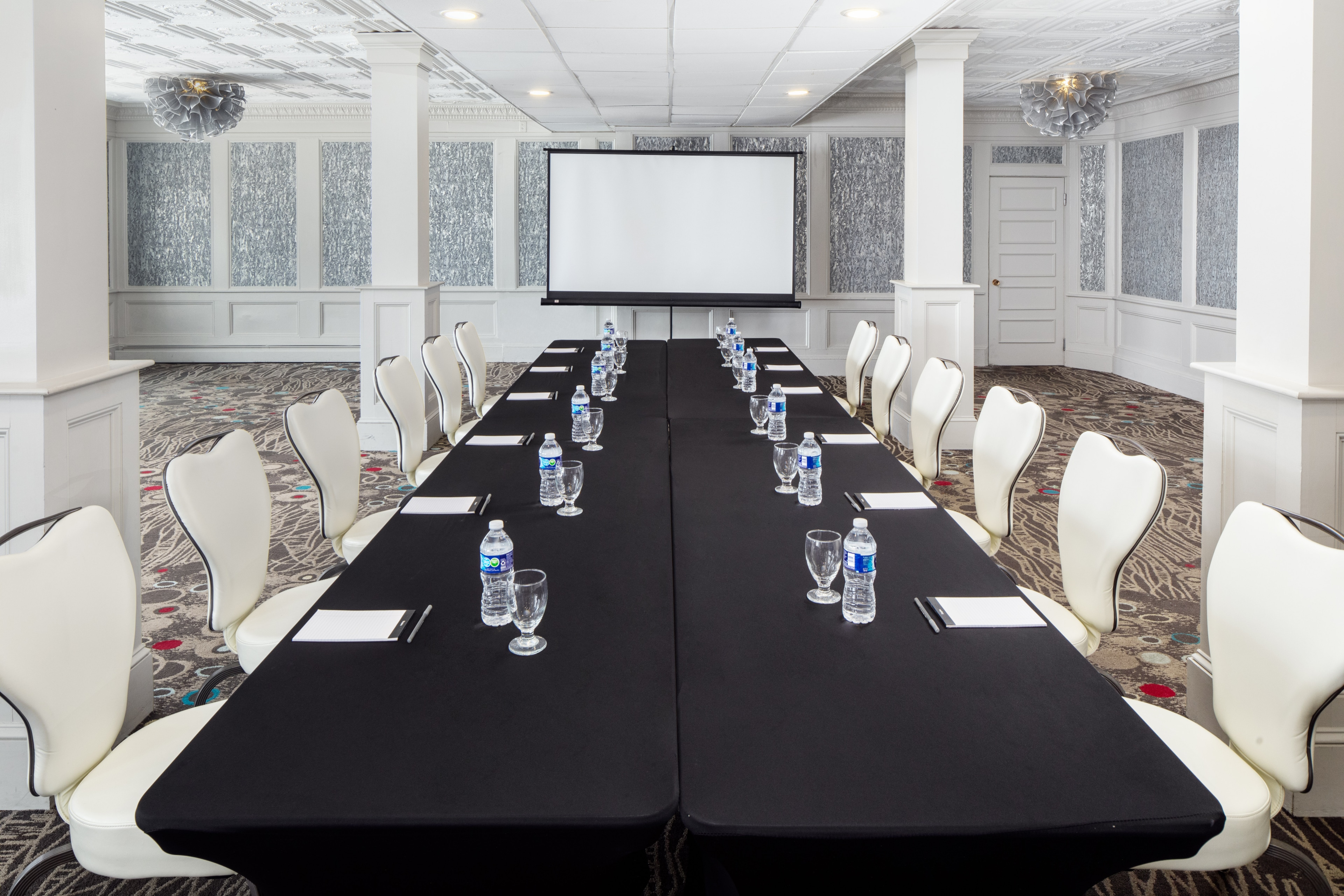 A meeting room with a long black table, white chairs, and a projector screen