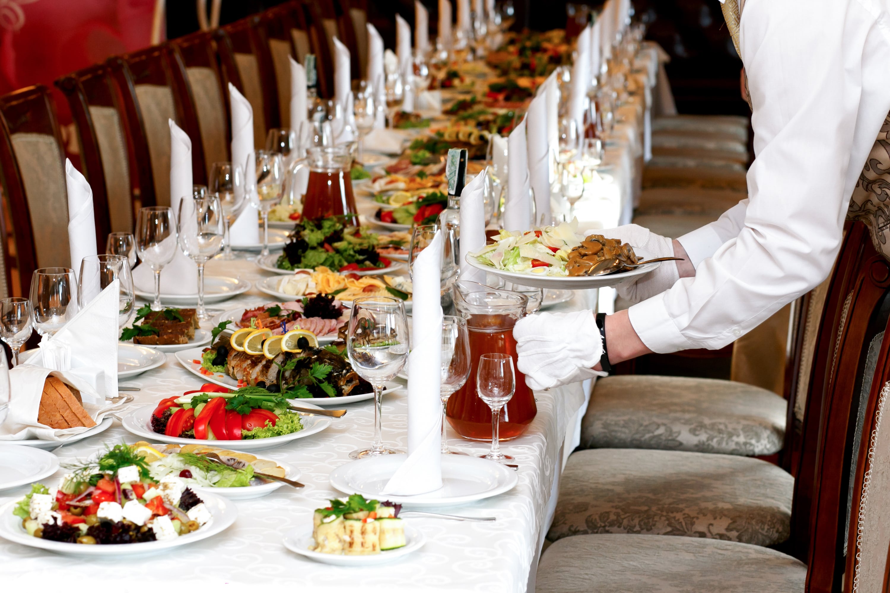 A formal dining table set with multiple dishes, wine glasses, and napkins, with a waiter serving food.