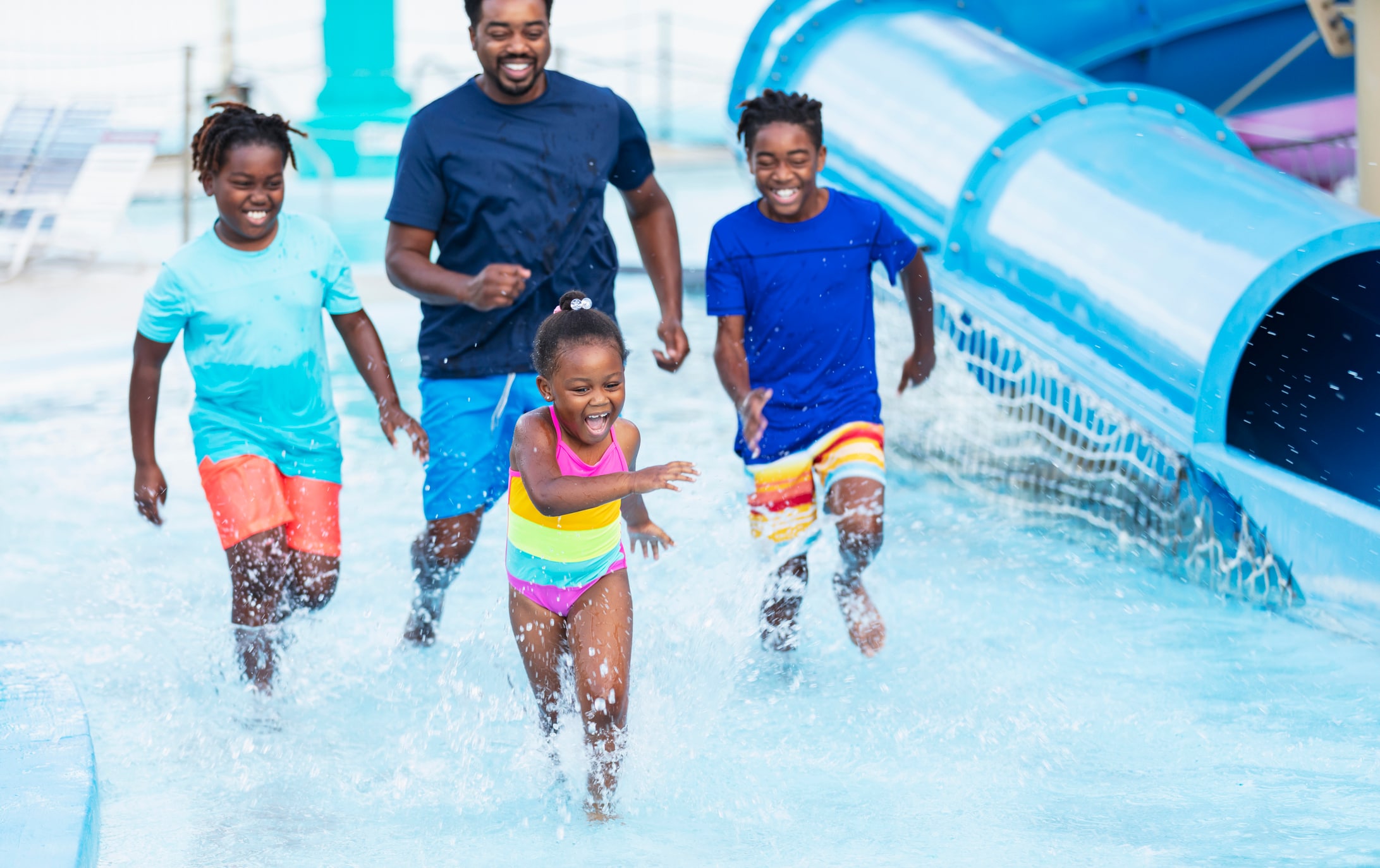 A family of four is running through a shallow pool area with a water slide.