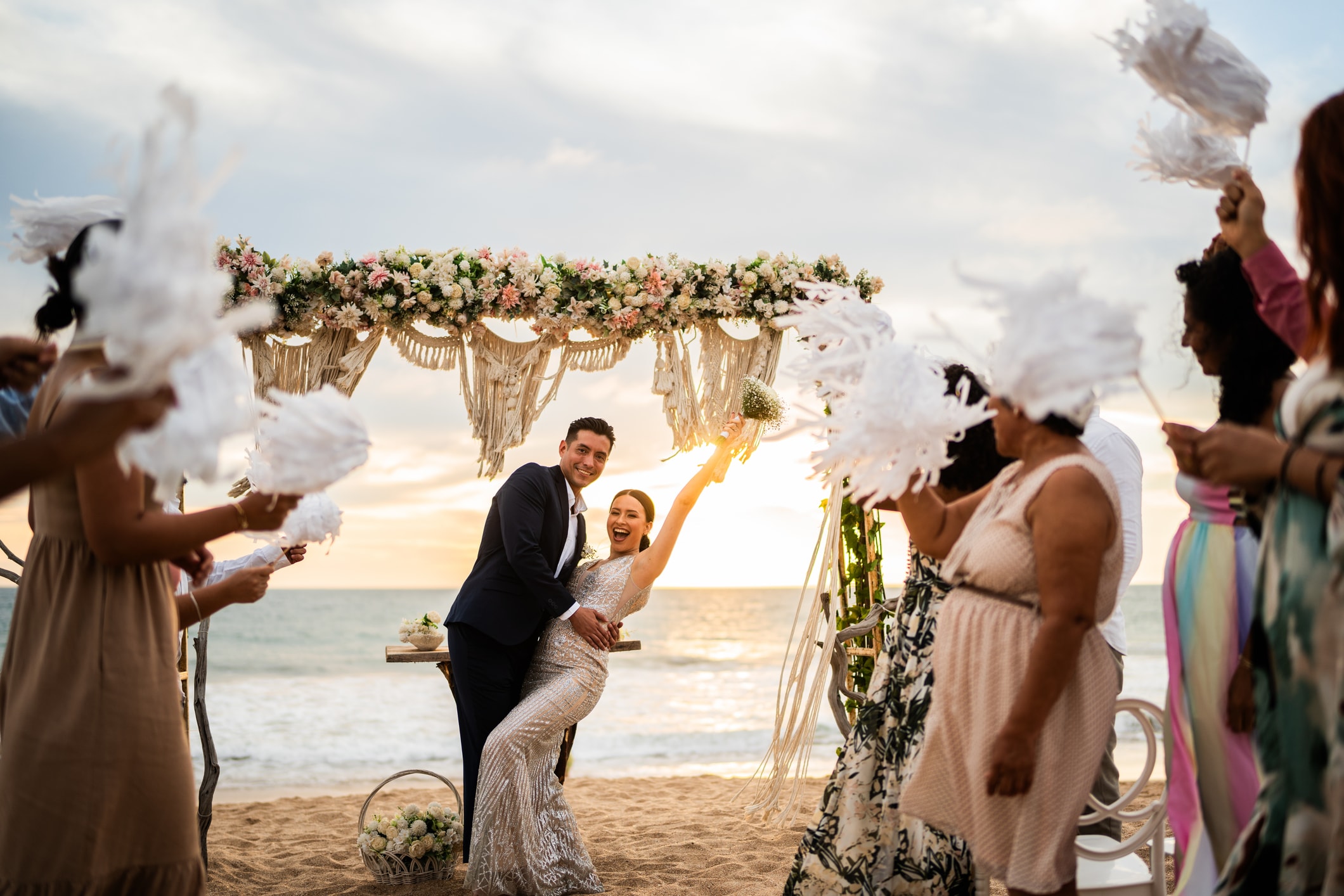 A beach wedding scene with a couple under a floral archway, surrounded by guests with feather fans.