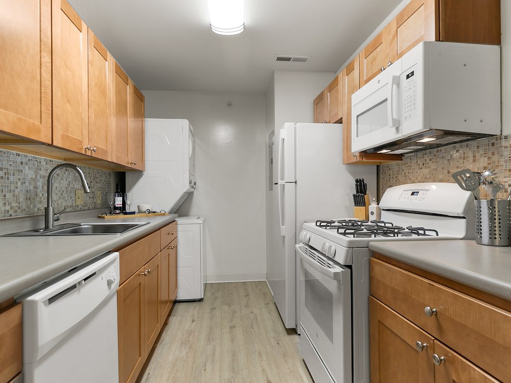 Contemporary Kitchen Style at California Apartments, shown here in the two-bedroom floor plan. Features warm maple wood cabinetry, white appliances, a tiled backsplash, and ample counter space.