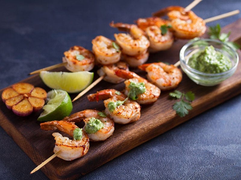 A wooden board with grilled shrimp skewers, a small bowl of green sauce, and lime wedges.