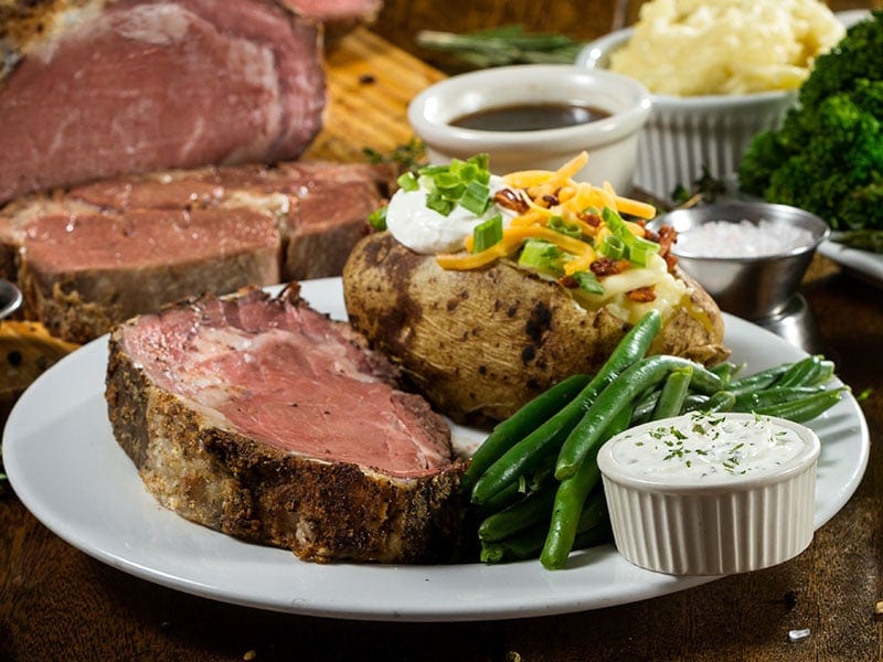 a plate of prime rib, a baked potato, green beans, and a small ramekin of sauce