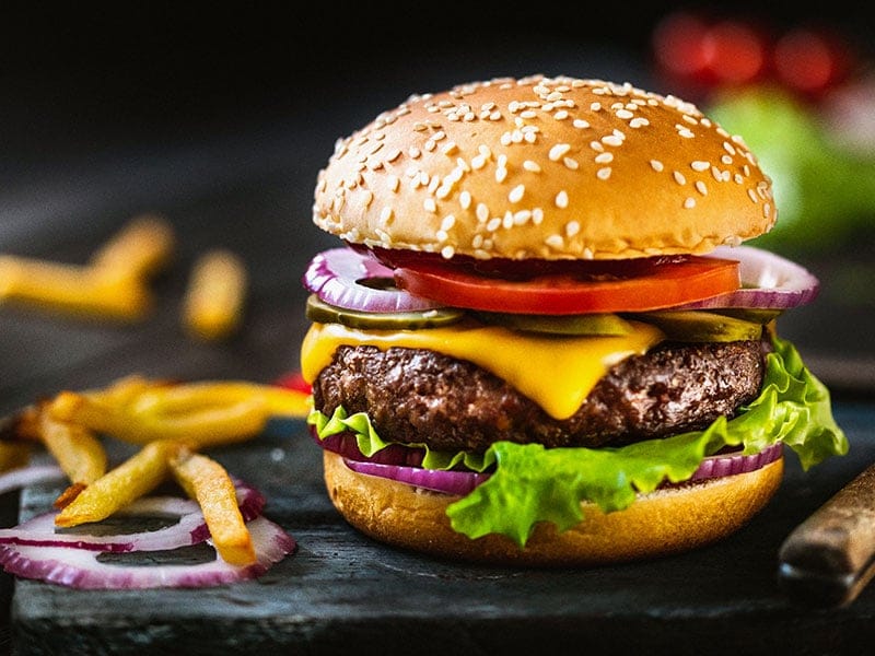 A cheeseburger with lettuce, tomato, onion, and cheese on a sesame seed bun with fries on the side.