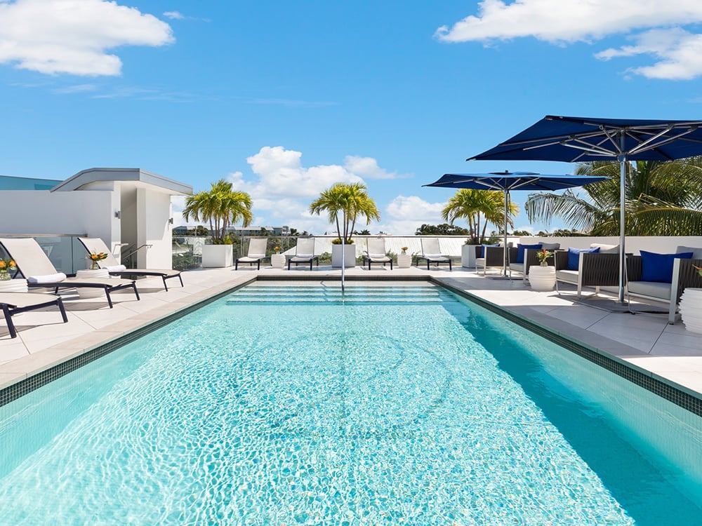A rectangular pool with clear blue water, surrounded by white lounge chairs and blue umbrellas, in front of a white building with palm trees.