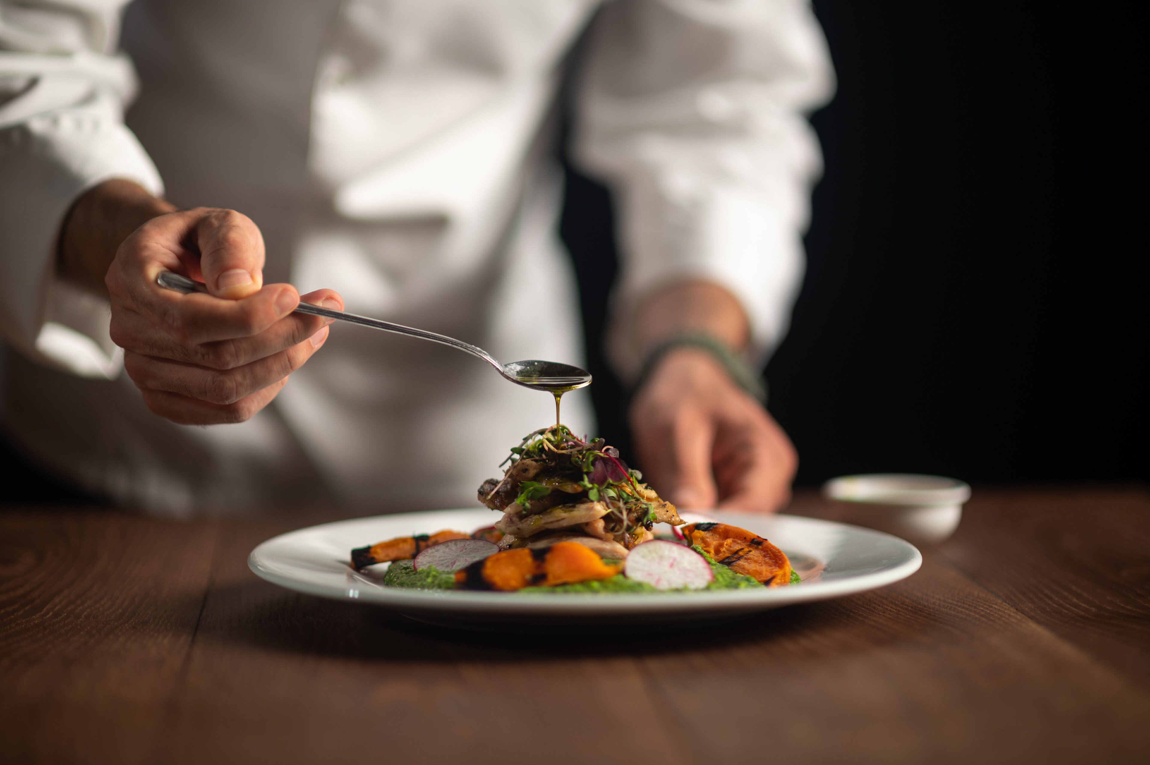 Chef drizzles sauce over a gourmet dish featuring vegetables, microgreens, and carefully arranged ingredients.